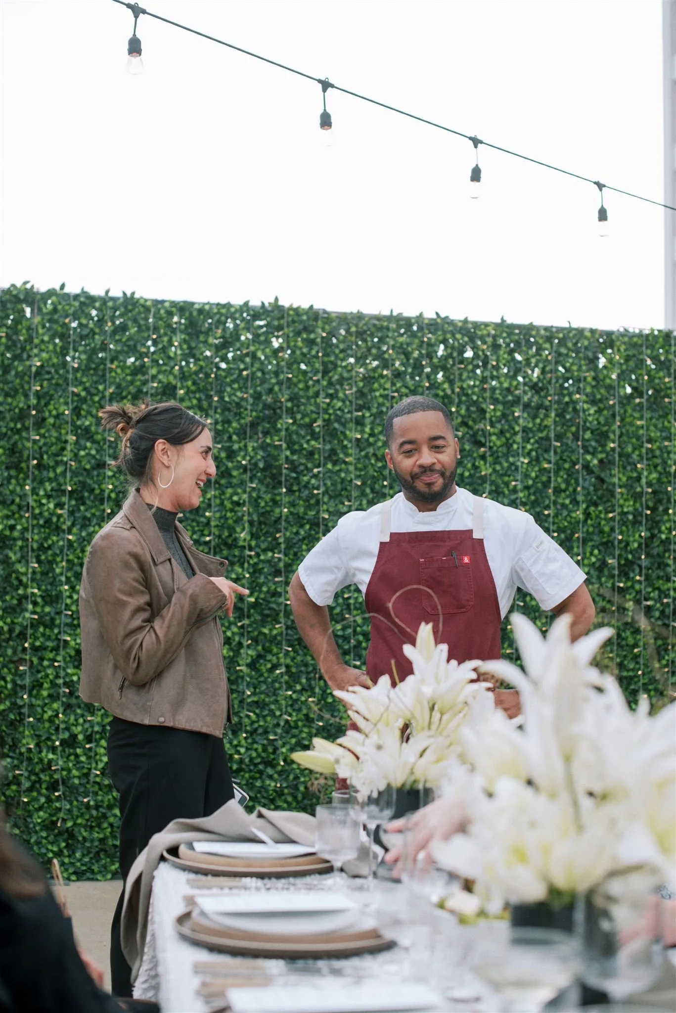Outdoor event space with elegant table setup, hedge backdrop, and string lighting