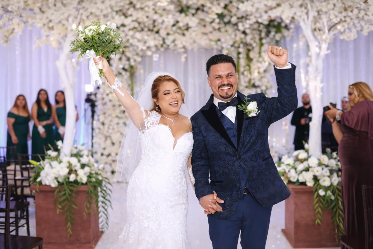 Bride and groom celebrate with raised arms in decorated chapel with white flowers and hanging greenery