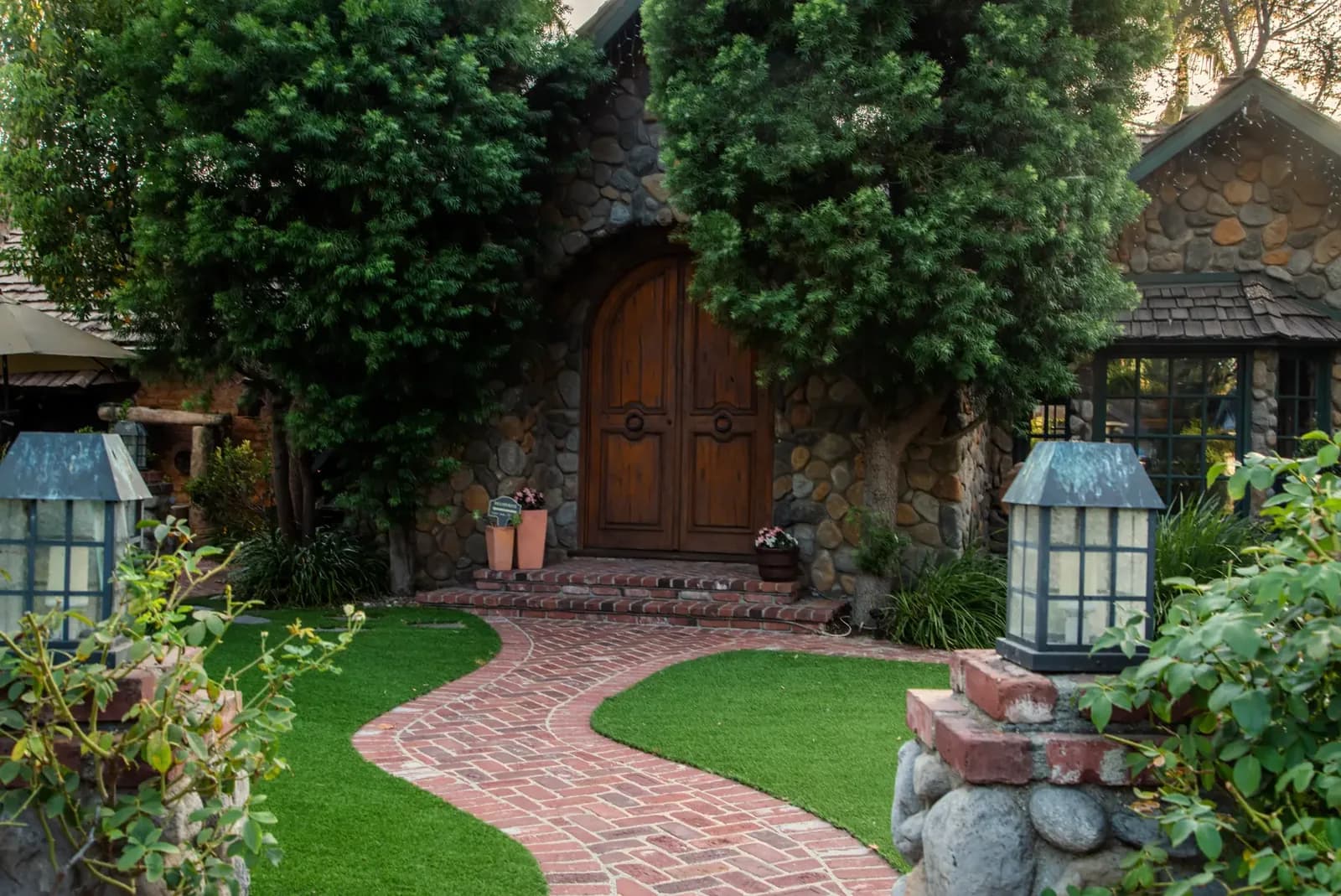 Stone cottage entrance with ivy, wooden doors, brick pathway, and green lawn with lanterns