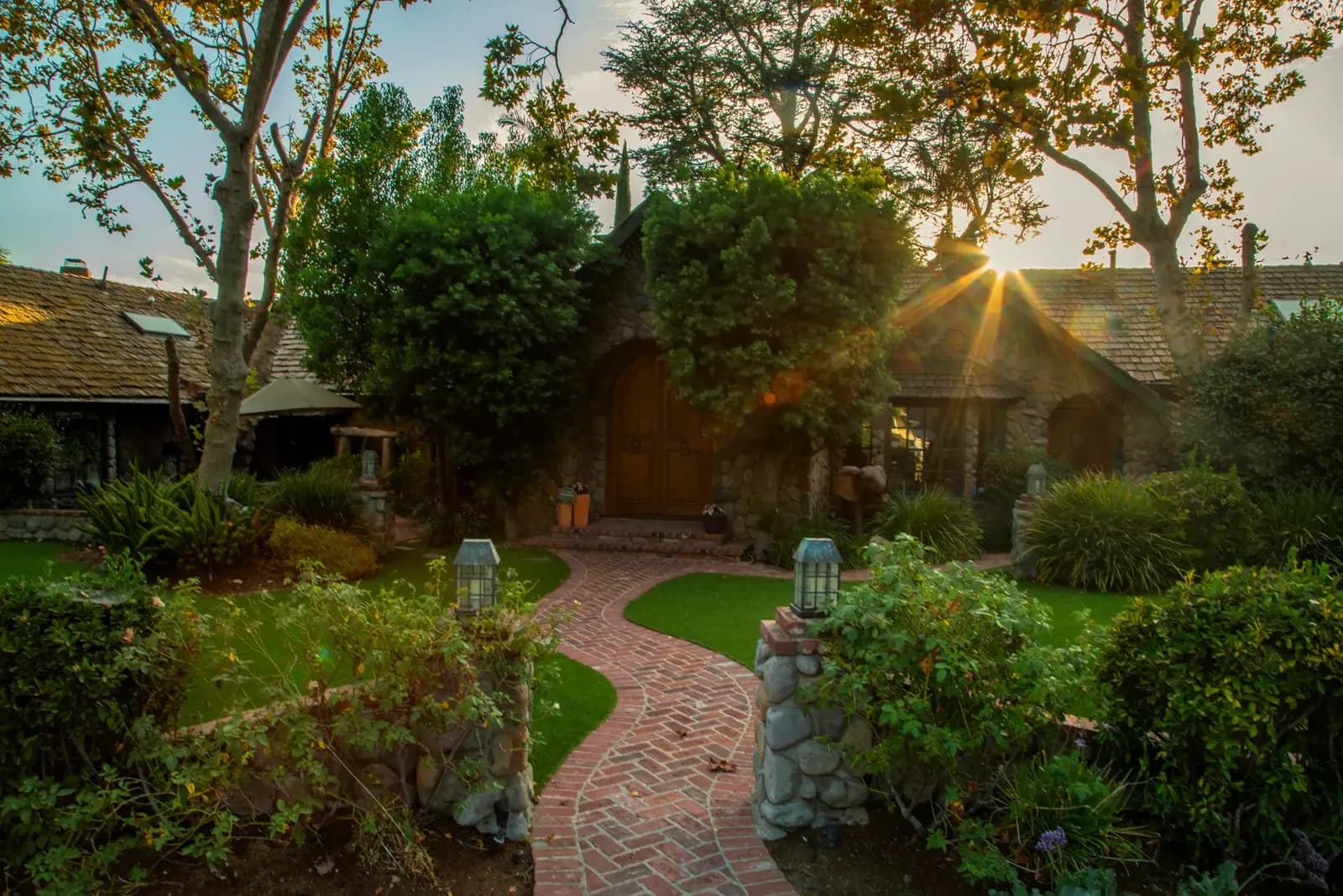 Enchanted Manor courtyard with curved brick path, green lawn, mature trees, and stone buildings at sunset