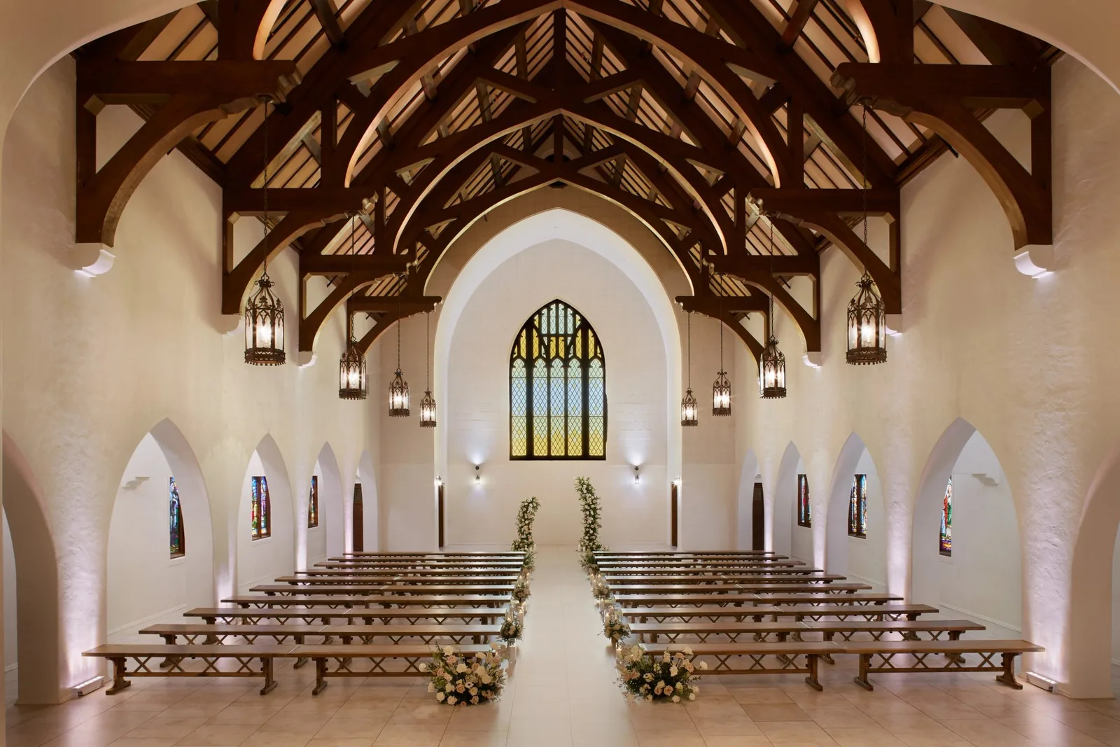 Wedding chapel interior with dramatic wooden beam ceiling, white walls, arched windows, and floral-lined aisle
