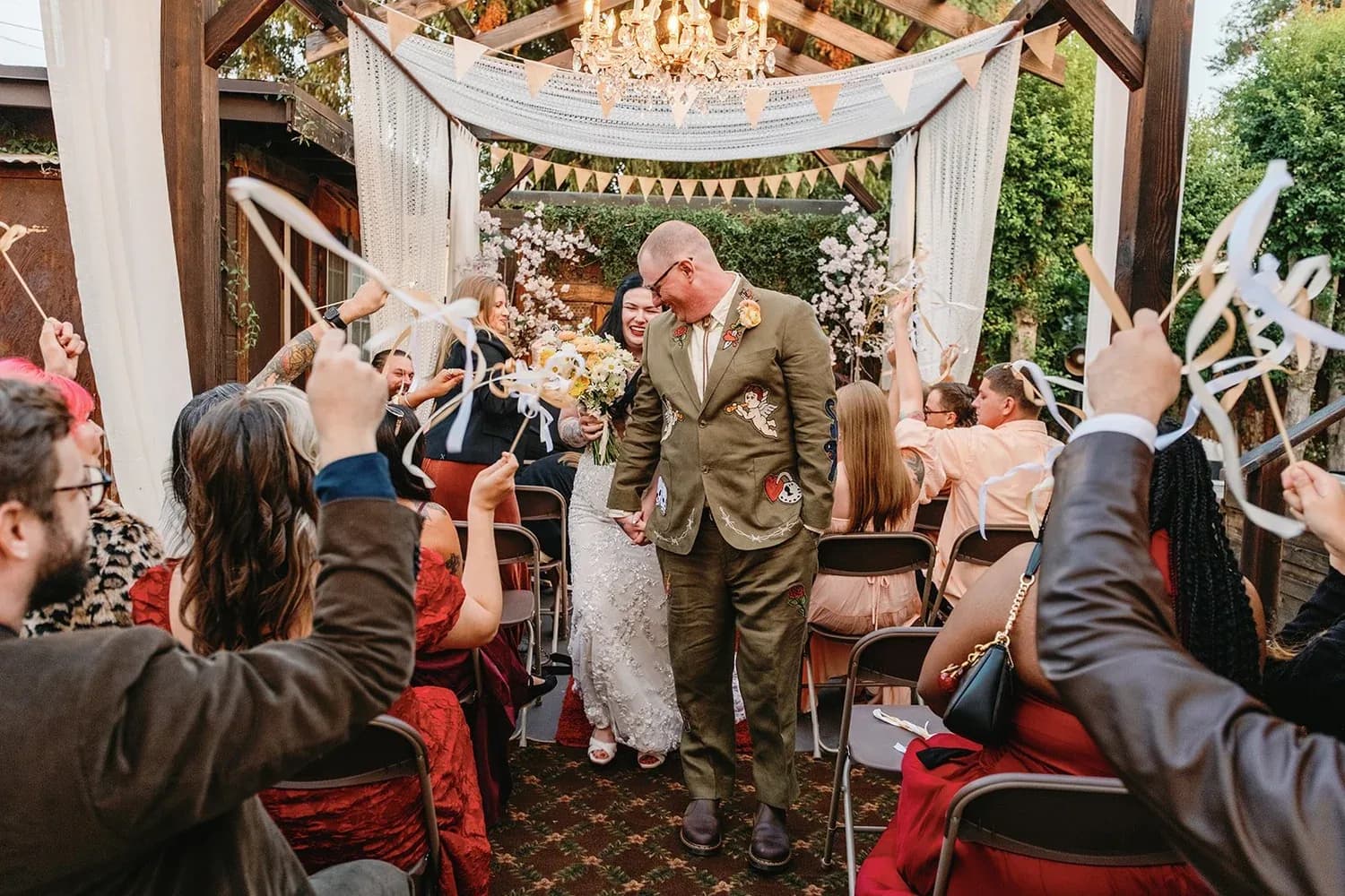 Bride and groom walking down aisle in decorated garden pavilion with cheering guests.