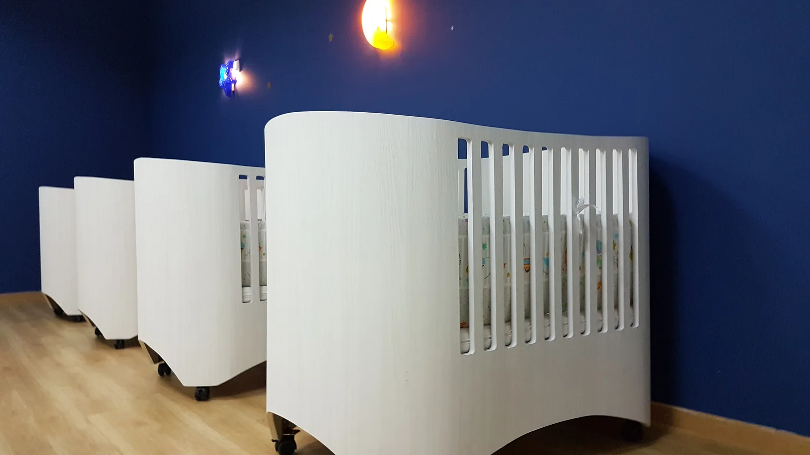 Multiple white curved baby cribs displayed in a preschool room with blue walls and wooden flooring.