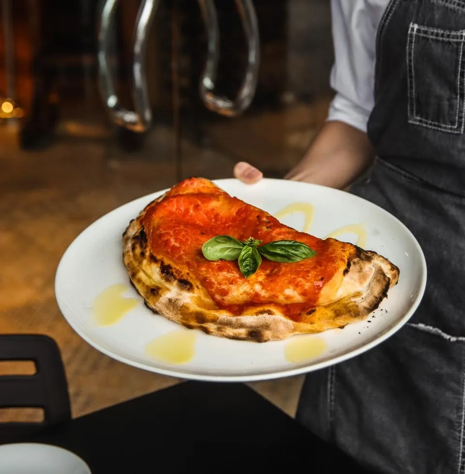 Wood-fired pizza with melted cheese and basil garnish on white plate, held by server in apron
