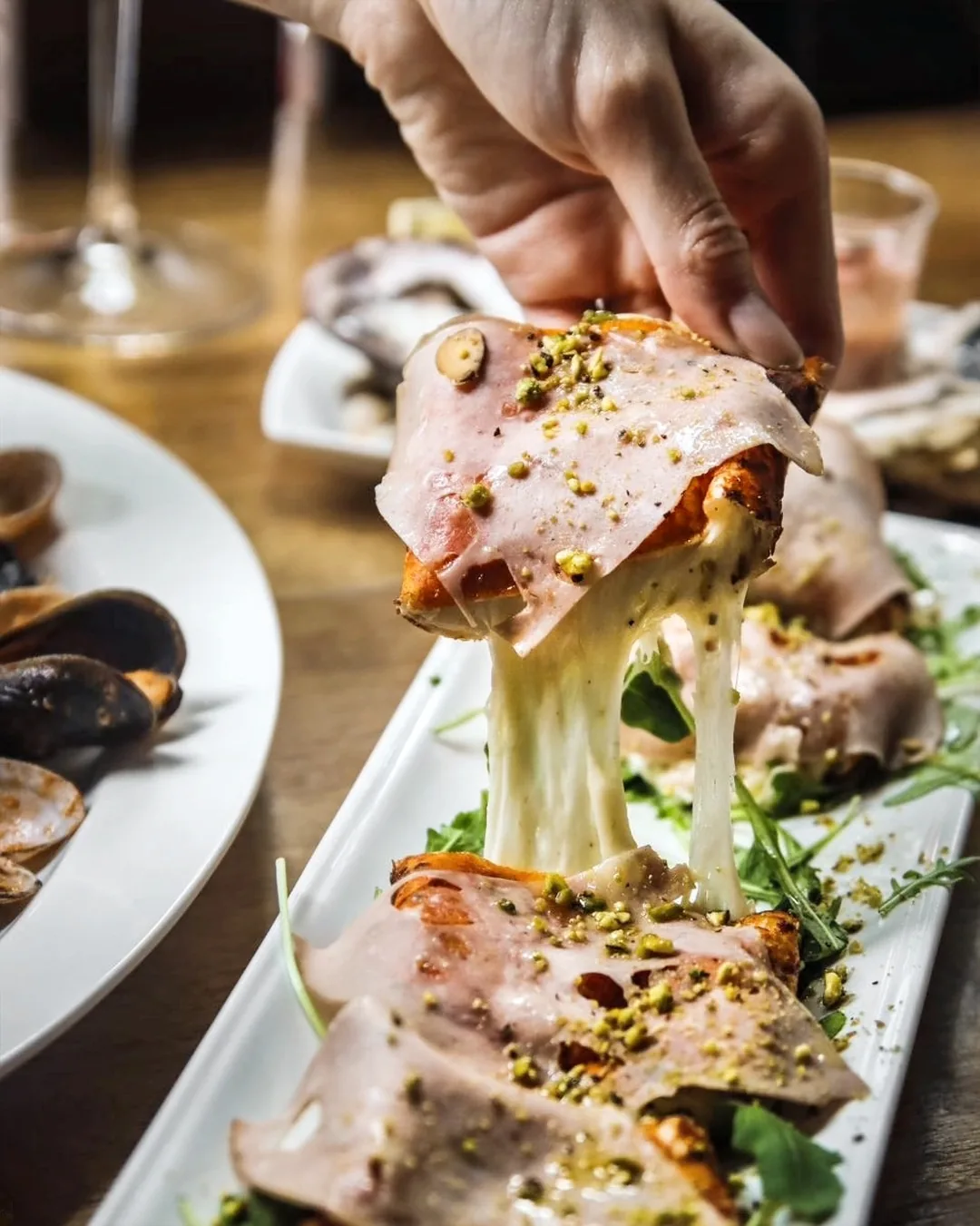 Hand lifting pizza slice with prosciutto and pistachio, melted cheese stretching