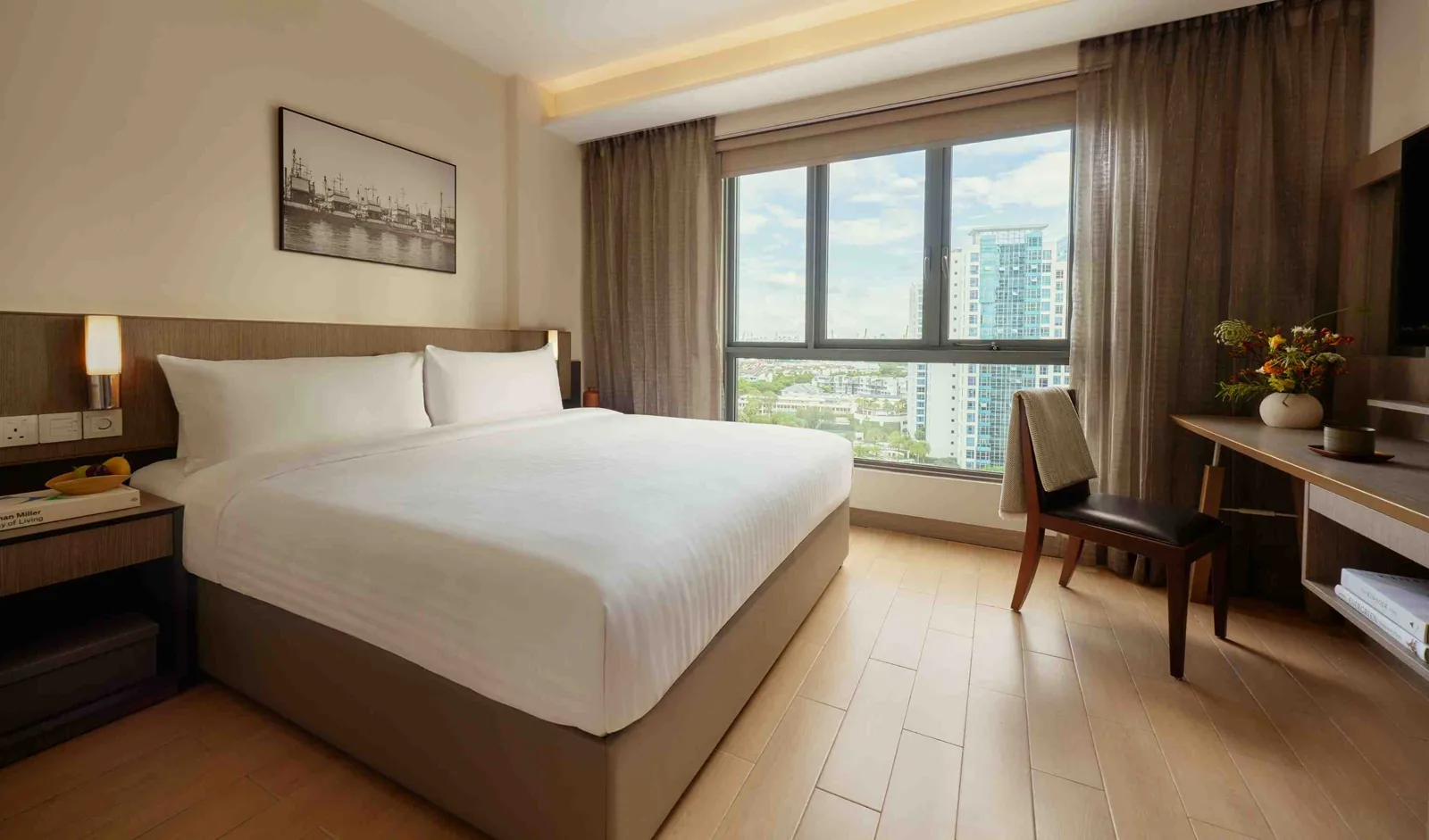 Modern hotel bedroom with white bedding, wooden desk, and city views through large windows