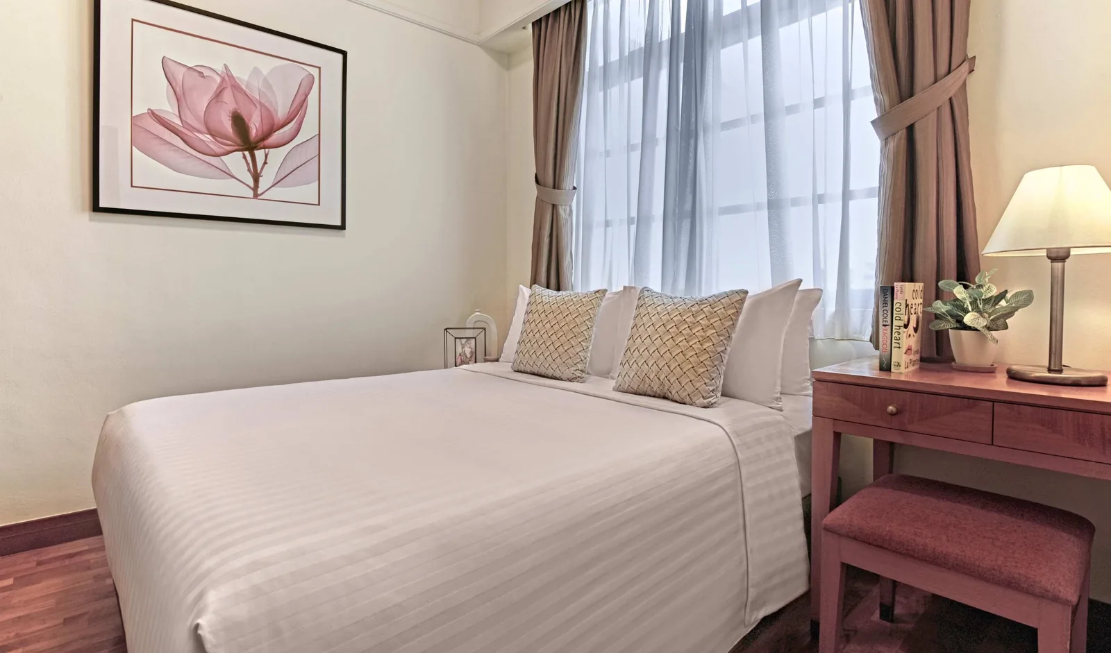 Hotel bedroom with white bed, burgundy desk, framed floral artwork, and large window with sheer curtains