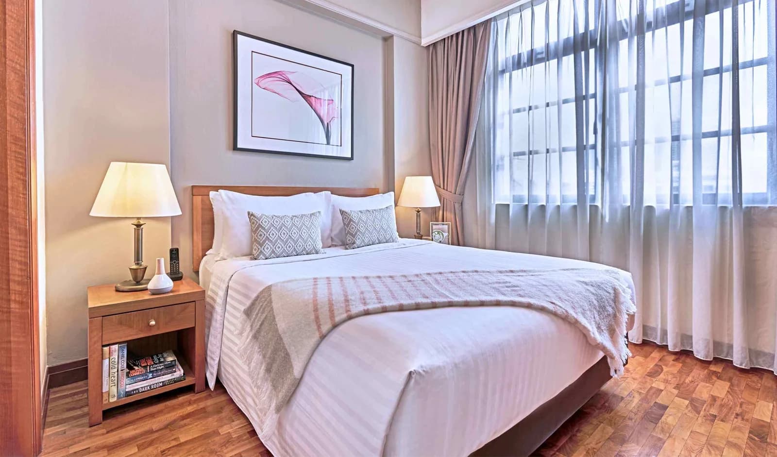 Bright hotel bedroom with queen bed, wooden furniture, and city views through large windows.