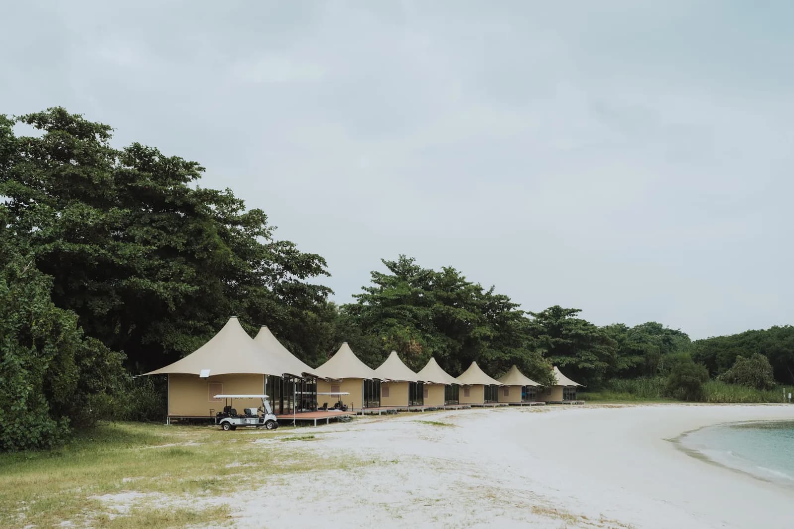 Beachfront glamping tents with white peaked roofs and a golf cart nestled among tropical trees