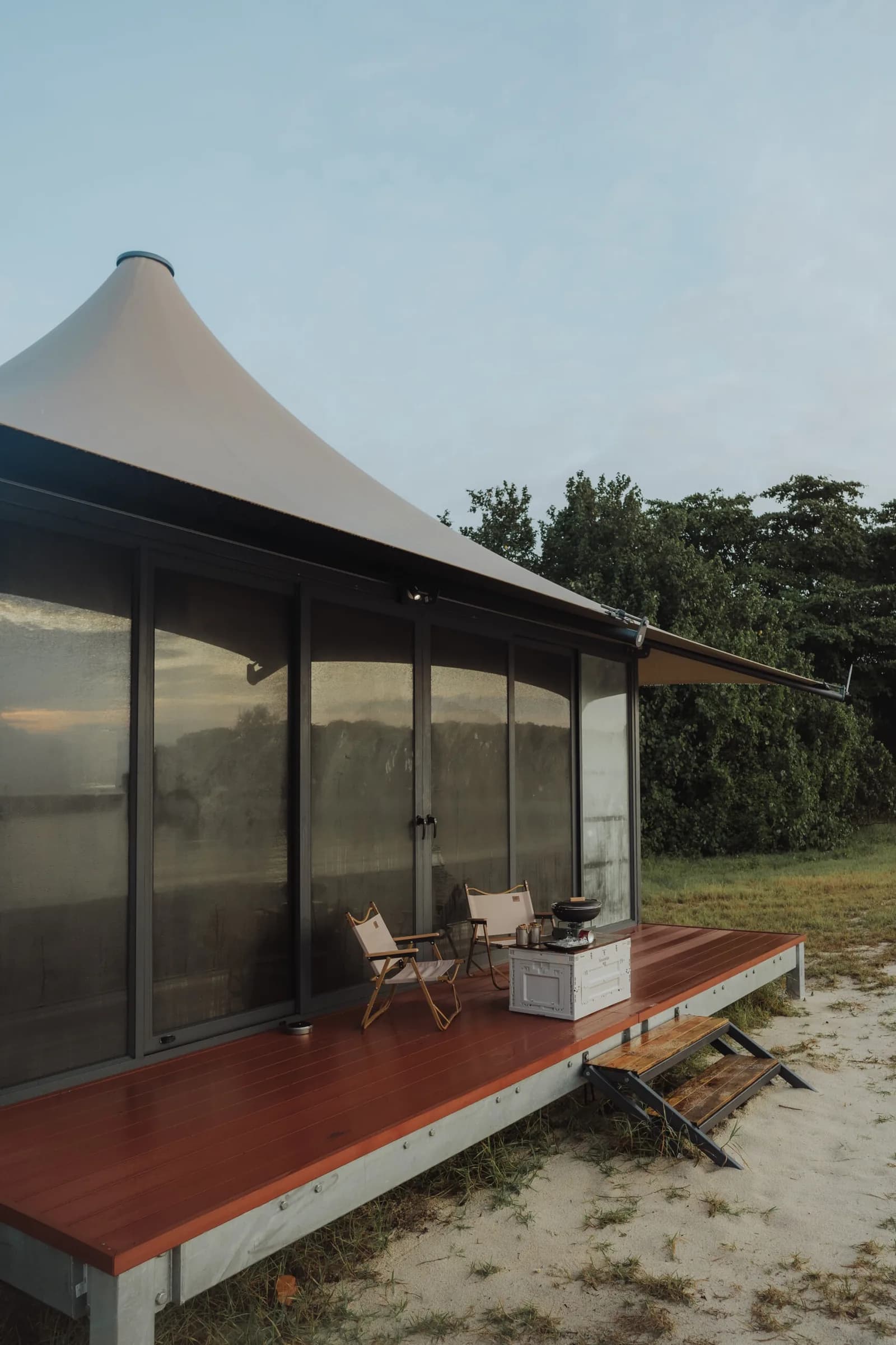 Modern glamping pavilion with white peaked roof and glass walls on wooden platform in natural setting