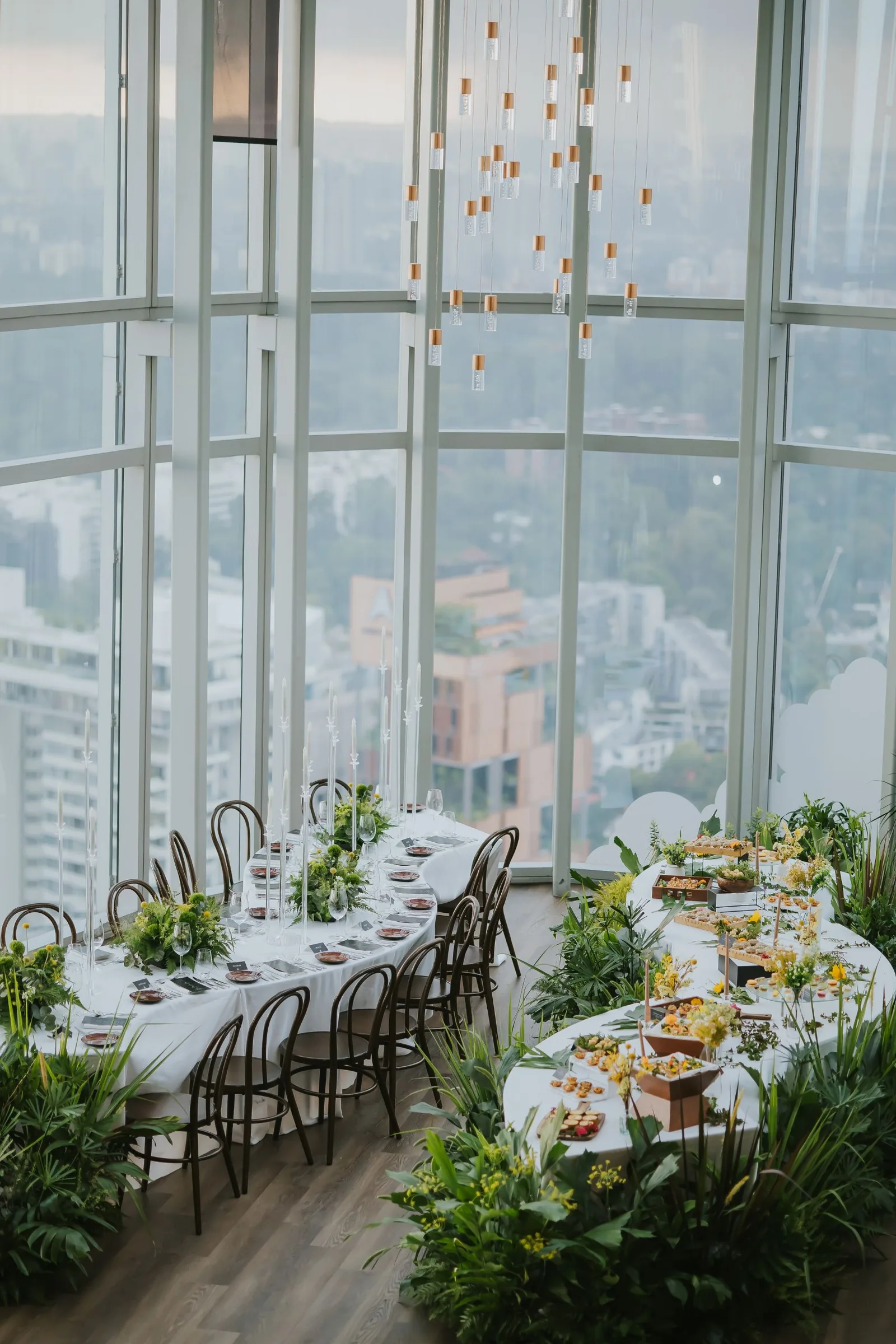 Elegant high-rise event space with panoramic city views, dressed tables with plants and pendant lighting