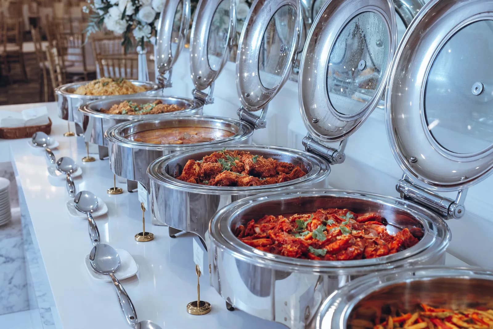 Buffet spread with multiple silver chafing dishes containing colorful curries and prepared dishes.