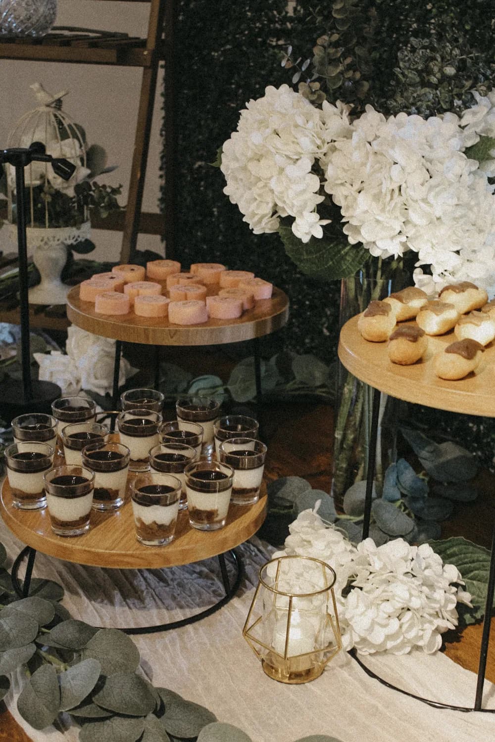 Tiered dessert display with layered treats, pastries, and coffee cups surrounded by white flowers and greenery.