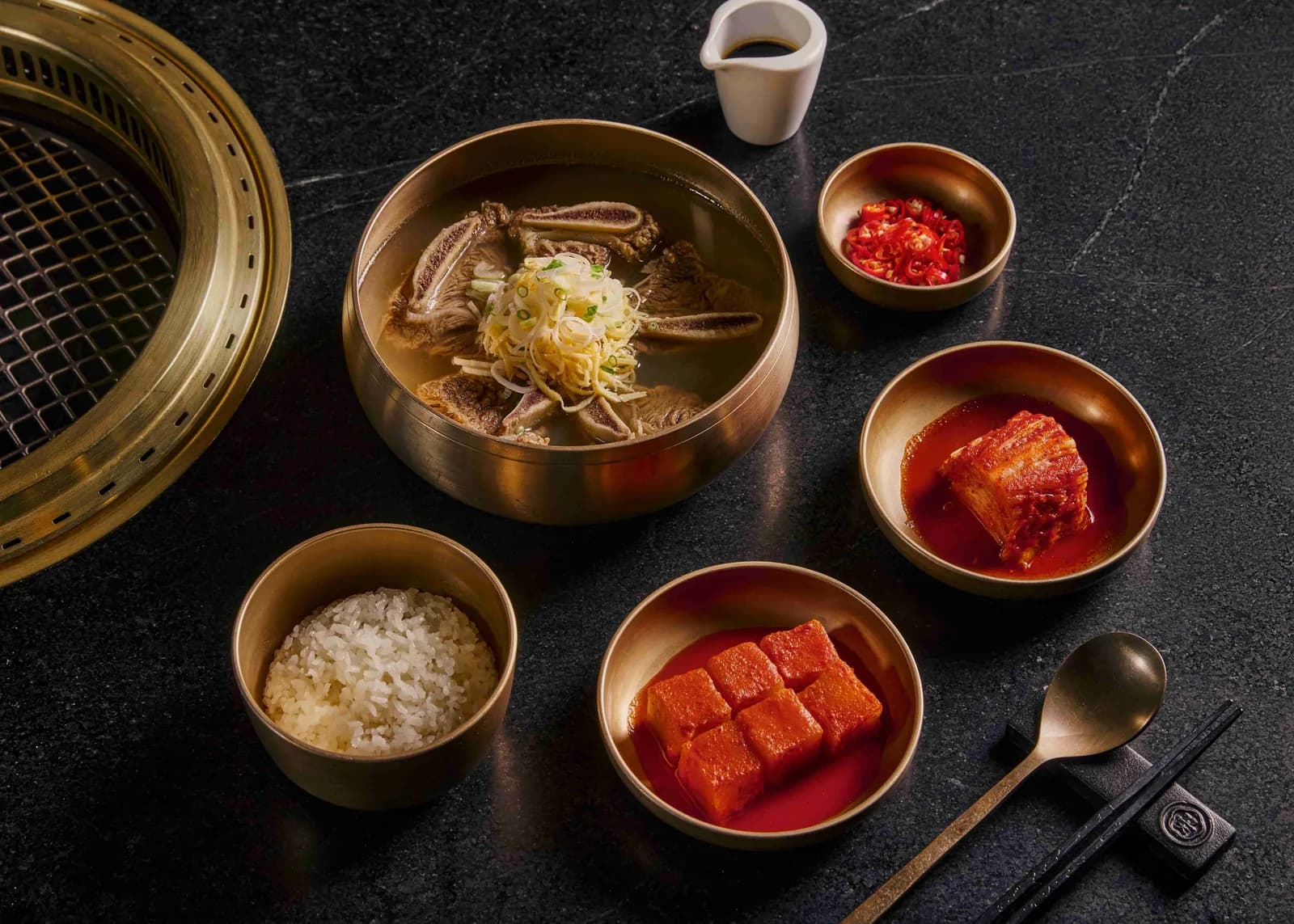 Korean BBQ ingredients and components arranged in brass bowls on a dark stone surface, including meat, vegetables, sauces, and steaming basket