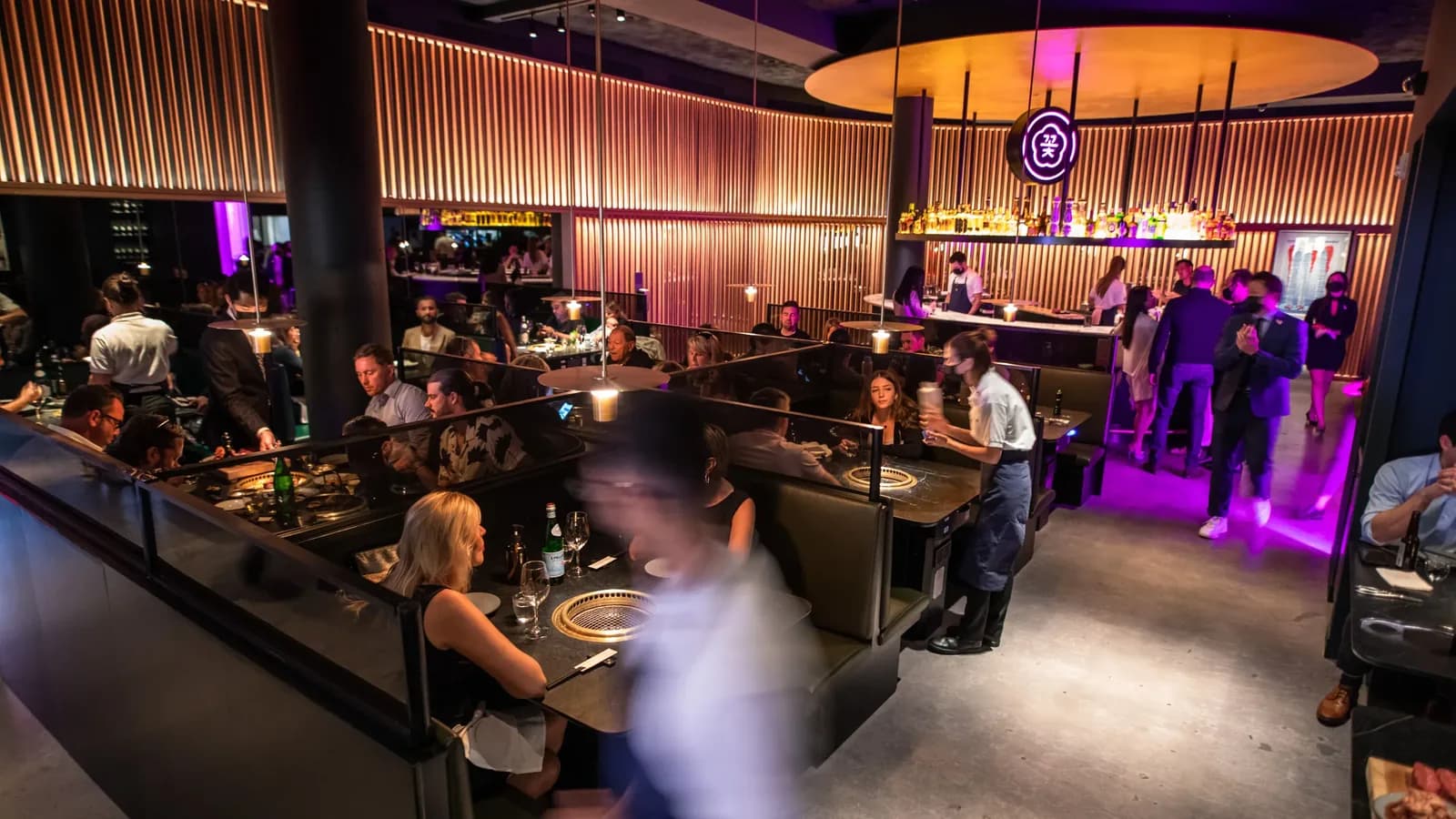 Modern restaurant interior with warm-lit wooden walls, purple lighting, and diners seated at tables throughout the space.