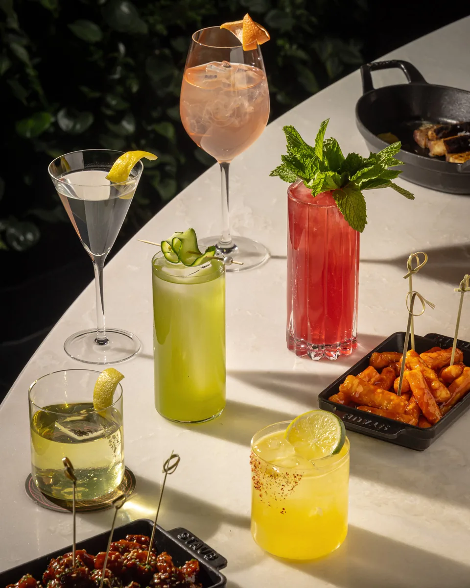Five colorful cocktails with citrus garnishes and fresh herbs, with appetizers on white table in sunlight