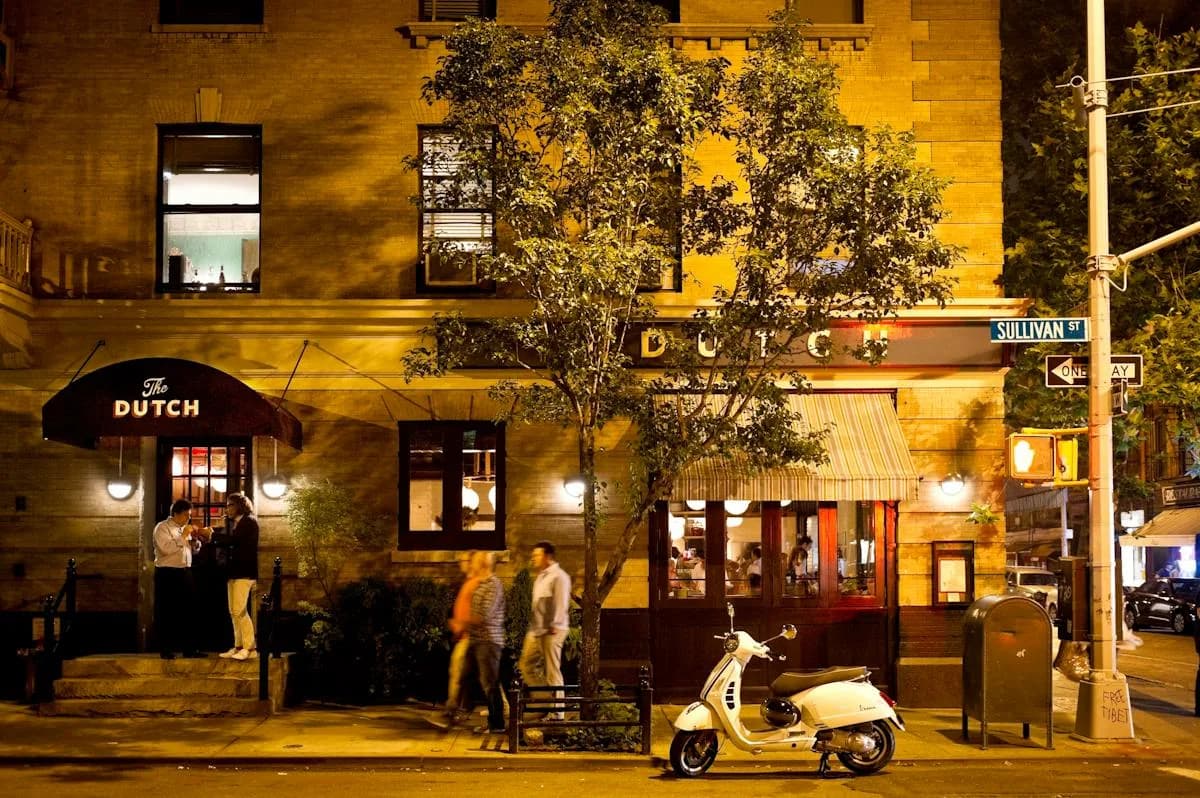 The Dutch restaurant storefront at night with black awning, lit windows, and ivy on yellow brick building