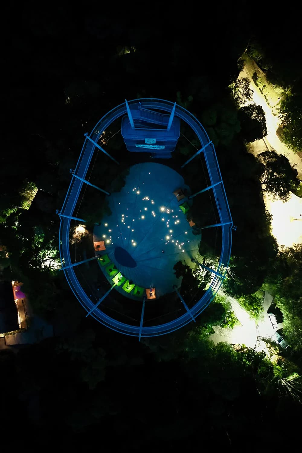 Lit canopy walkway at night viewed from above, with blue railings and tropical surroundings.