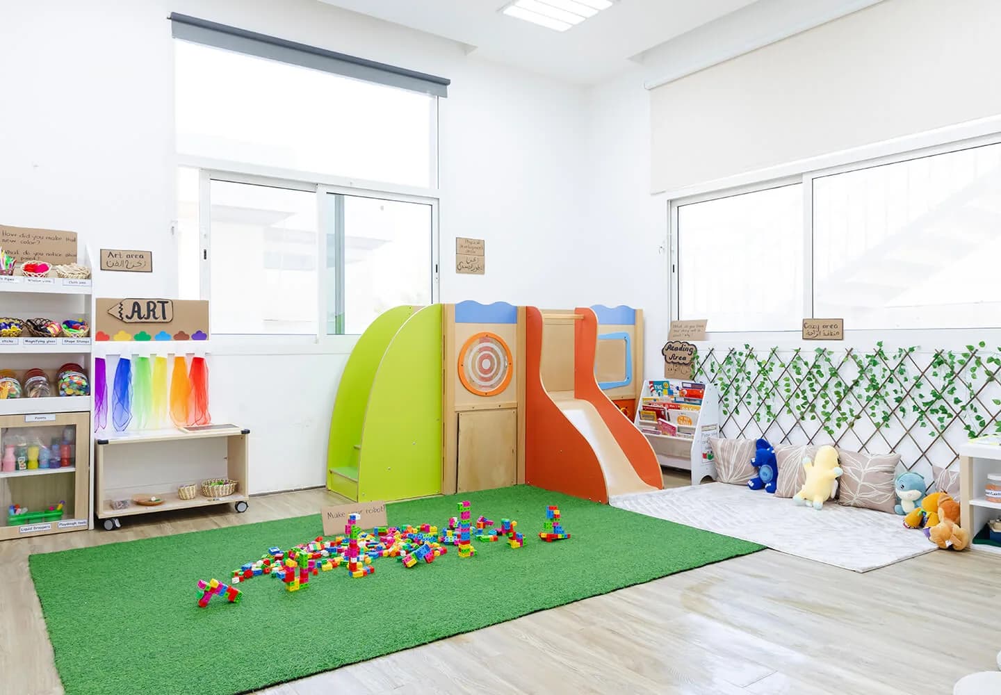 Bright nursery classroom with colorful slide structure, shelving with supplies, and toys on green mat