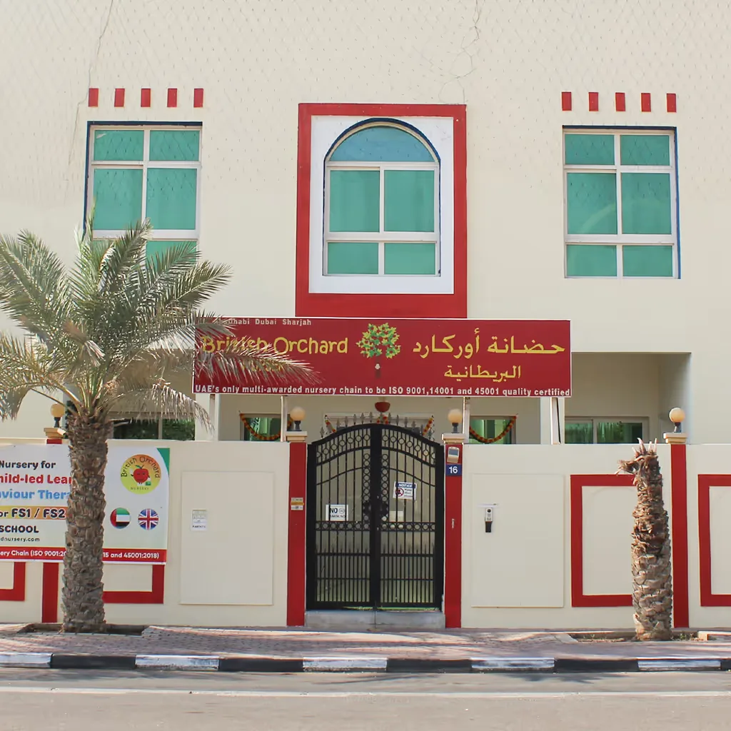 Red and white nursery building with arched windows, palm trees, and black metal entrance gate