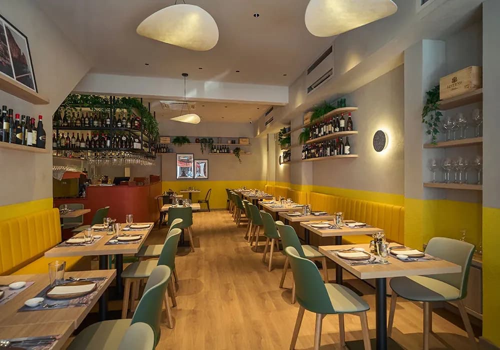 Bright dining room with yellow booth seating, wooden tables, green chairs, and shelves of wine bottles behind the bar