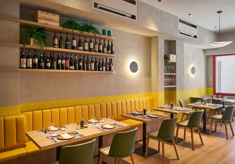 Modern restaurant dining room with yellow banquette seating, wooden tables, and wine bottles displayed on wall shelves