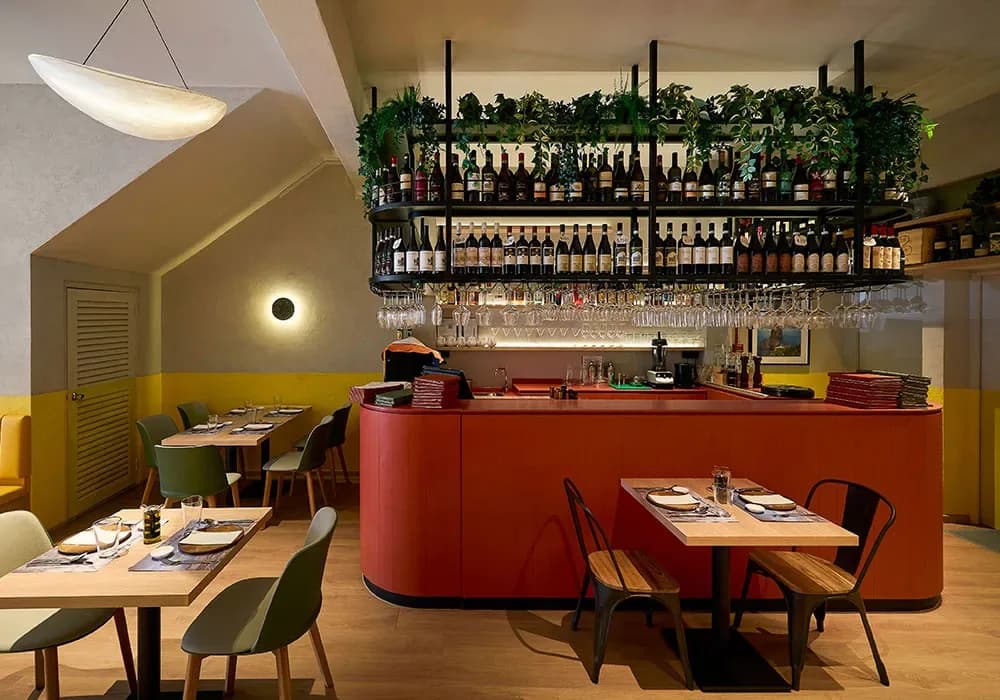 Modern restaurant interior with yellow walls, red bar counter, plant-topped shelving, and dining tables set for service.