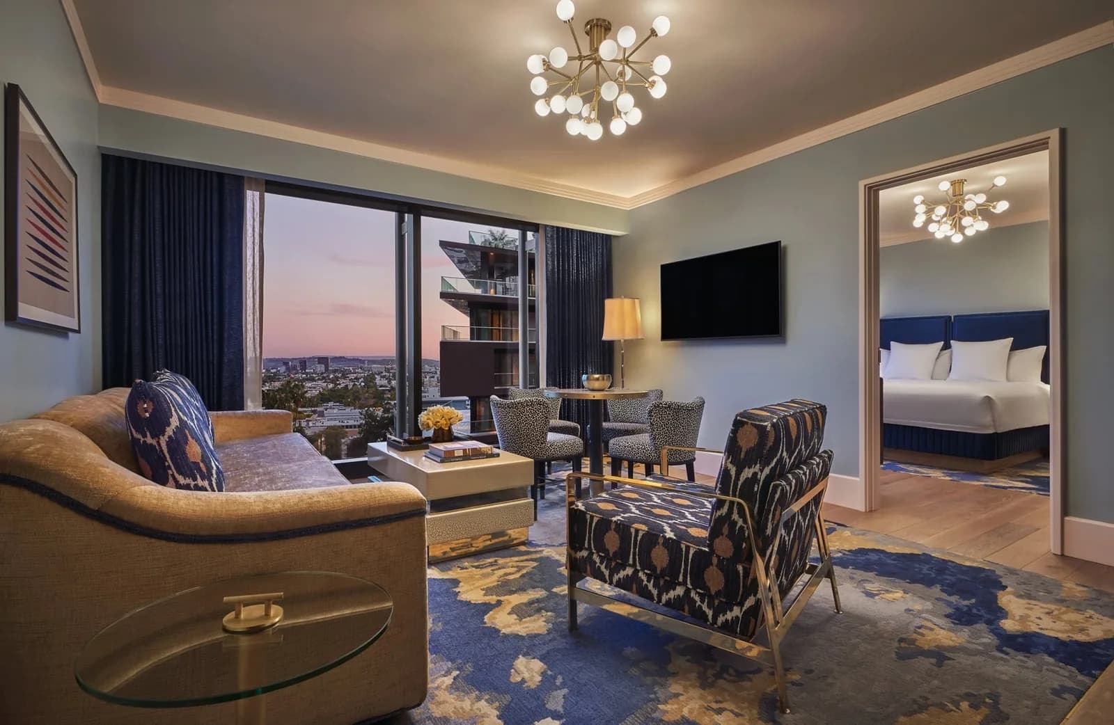 Upscale hotel suite with curved gold sofa, patterned chairs, and panoramic city skyline view through floor-to-ceiling windows at dusk.