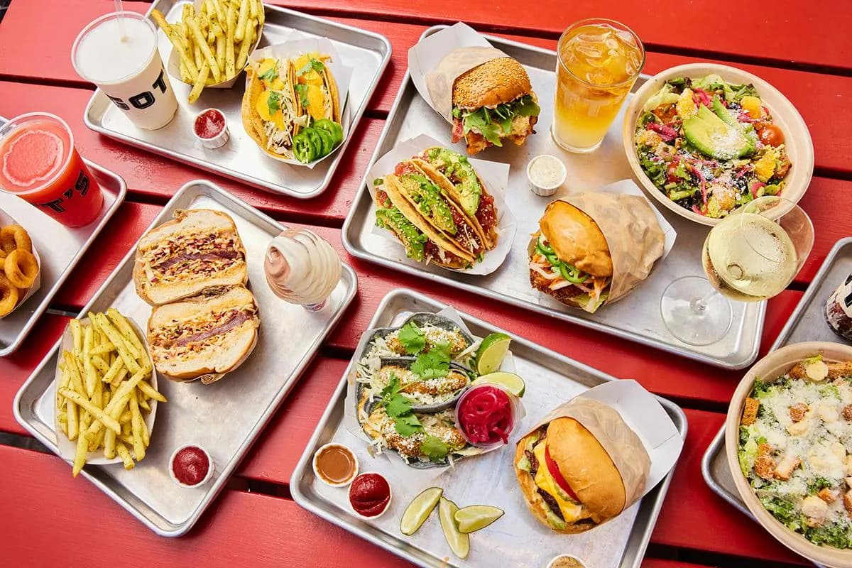 Overhead view of multiple dishes including tacos, burgers, fries, salads, and drinks on white trays on a red table