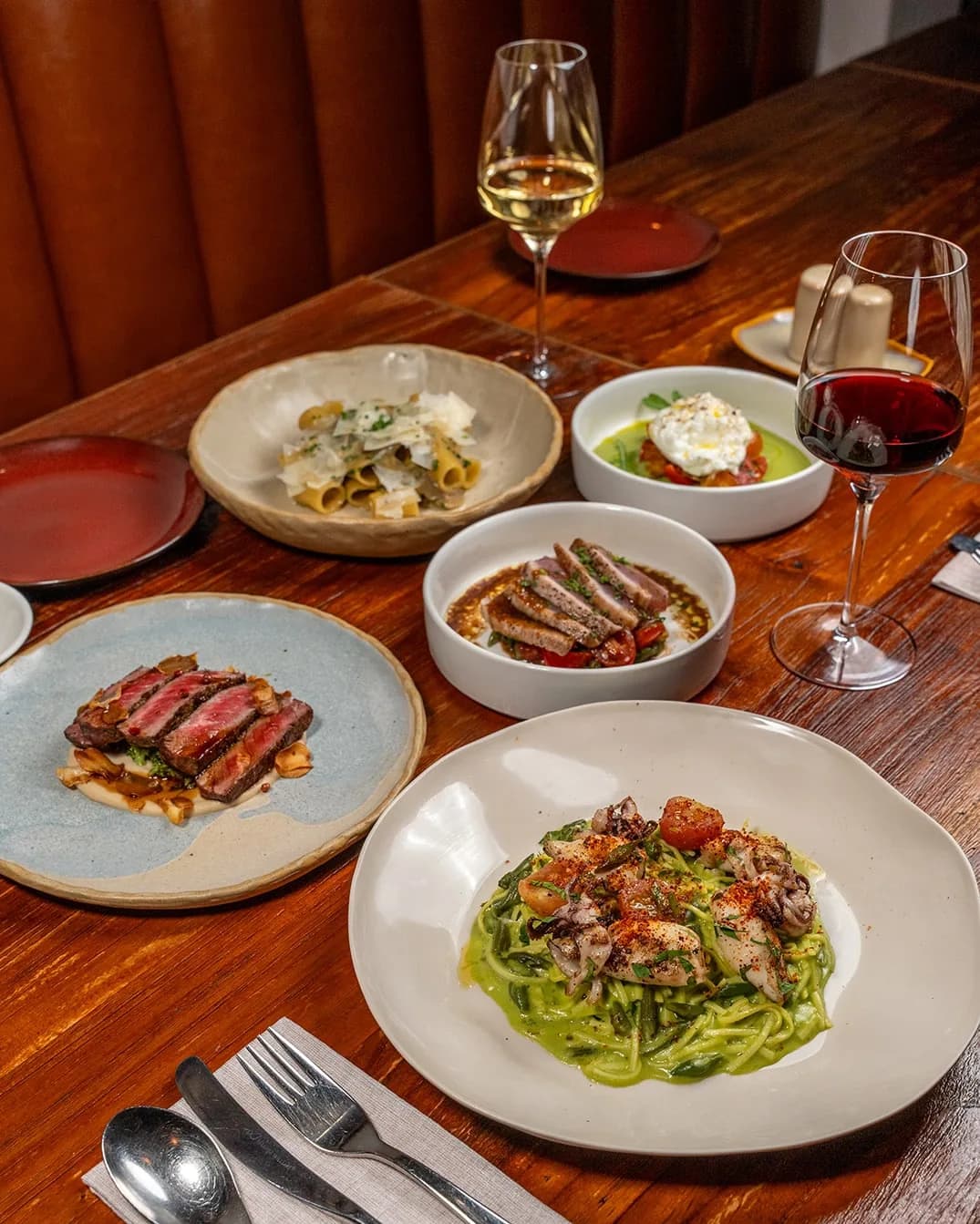 Fine dining table with plated steak, green pasta, and seafood dishes with wine glasses