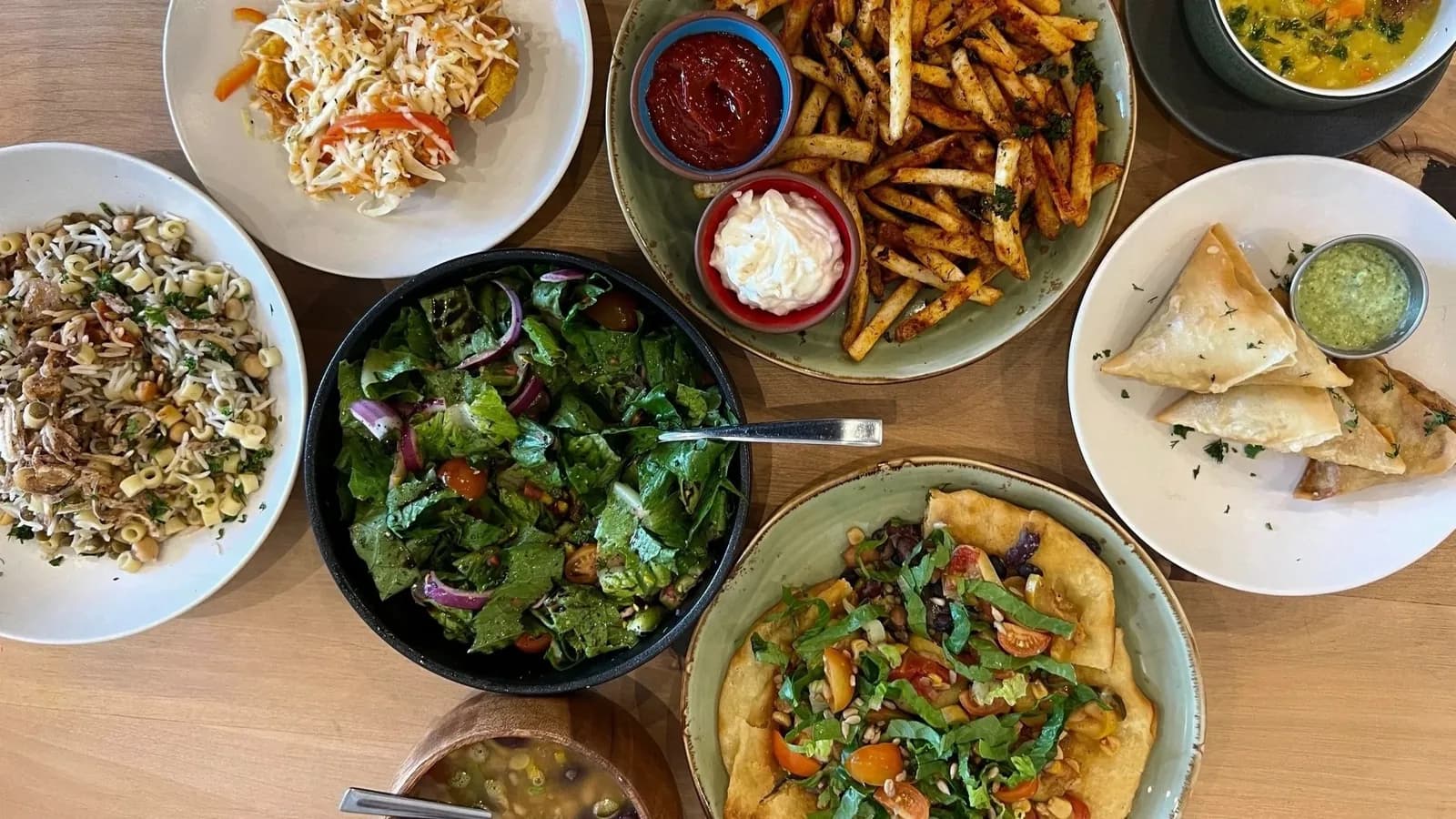 Restaurant spread with salads, fries, pastries, flatbread, and dipping sauces on wooden table