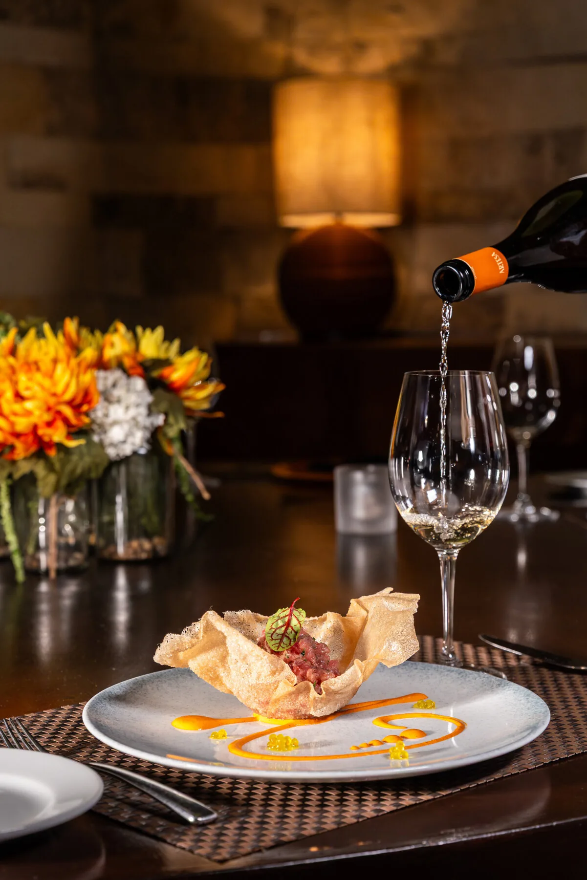 Wine being poured over a fine dining plate with crispy pastry appetizer and floral arrangement