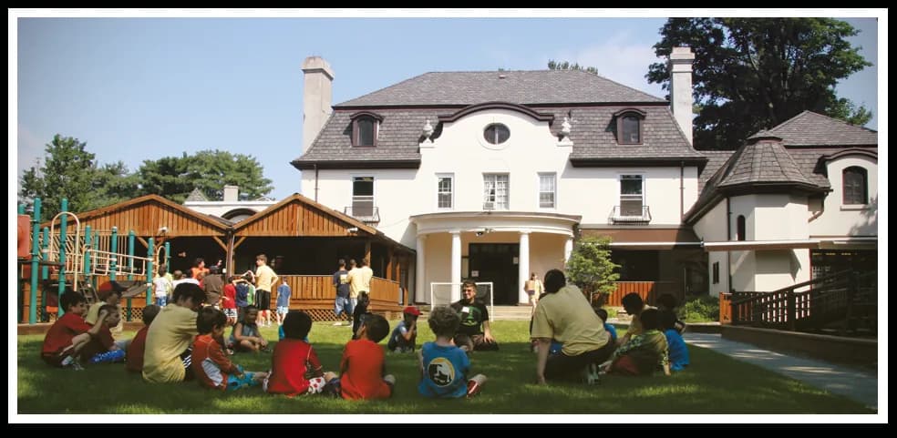 Children playing on the lawn in front of a large white mansion with wooden play equipment and porches.