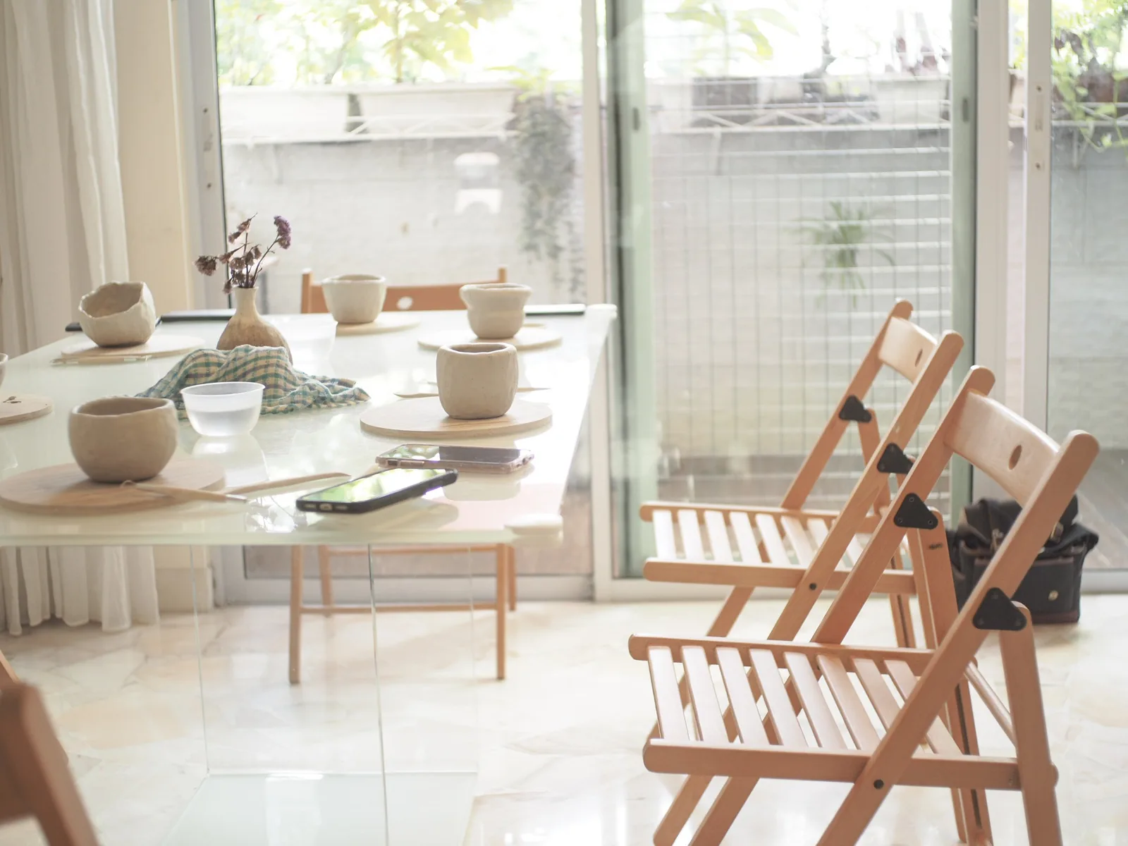 Pottery studio interior with ceramic bowls and vessels on display table, wooden chairs, and bright windows.
