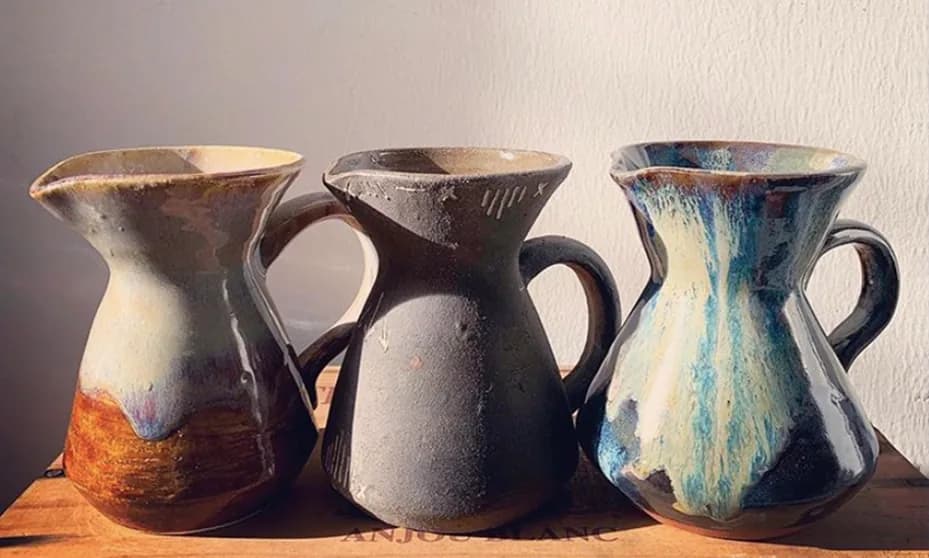 Three ceramic pitchers with varied glazes and hand-formed details displayed on a wooden surface