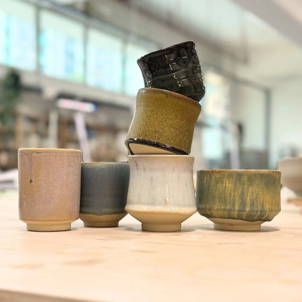 Stacked handmade ceramic cups and vessels with earth-toned and dark glazes in a pottery studio