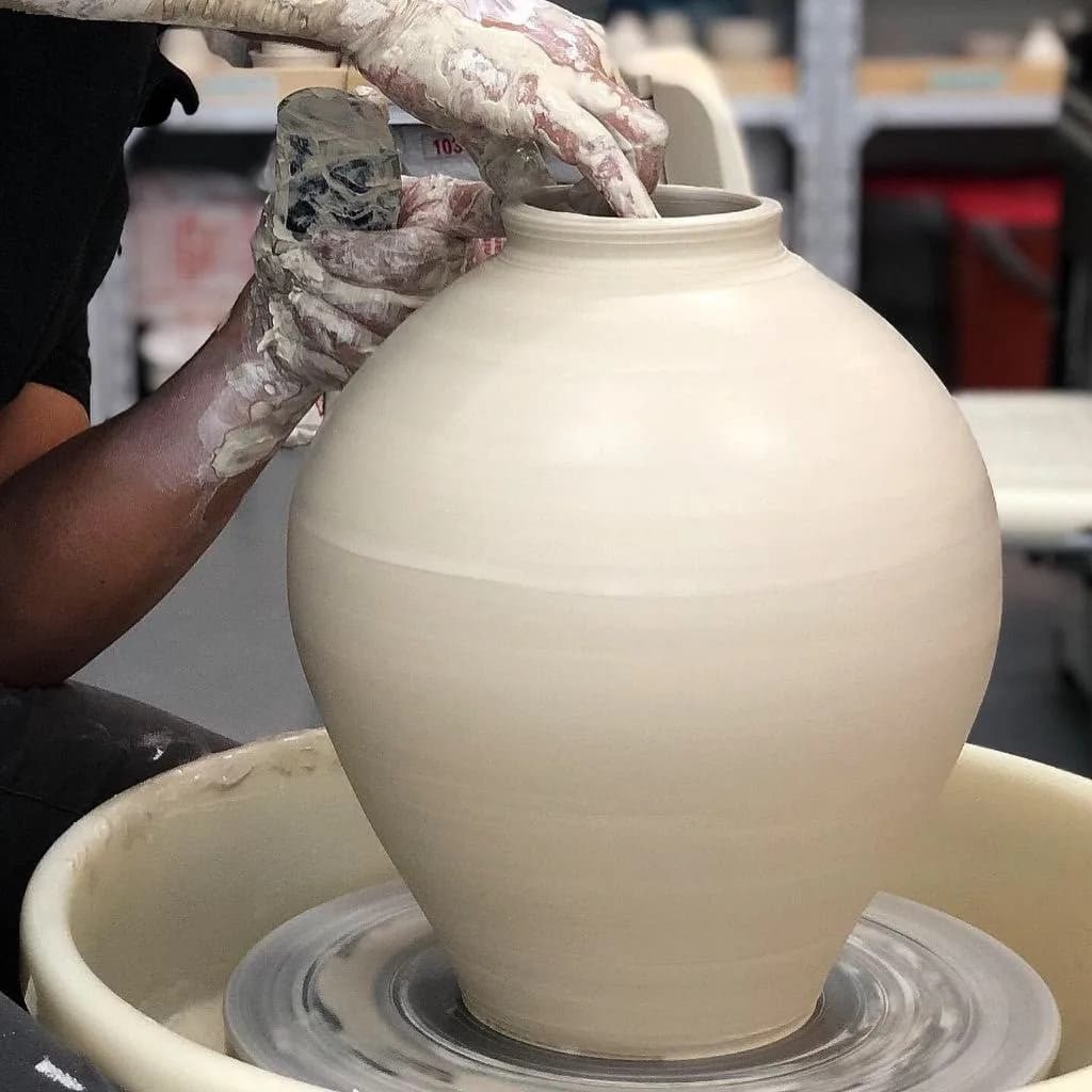 Potter's clay-covered hands shaping a large rounded ceramic vessel on a spinning wheel