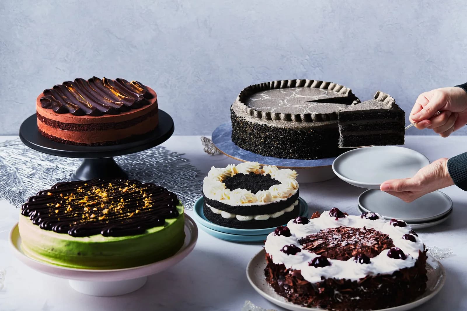 Five decorated cakes displayed with one being served, including chocolate, matcha, and white cream varieties