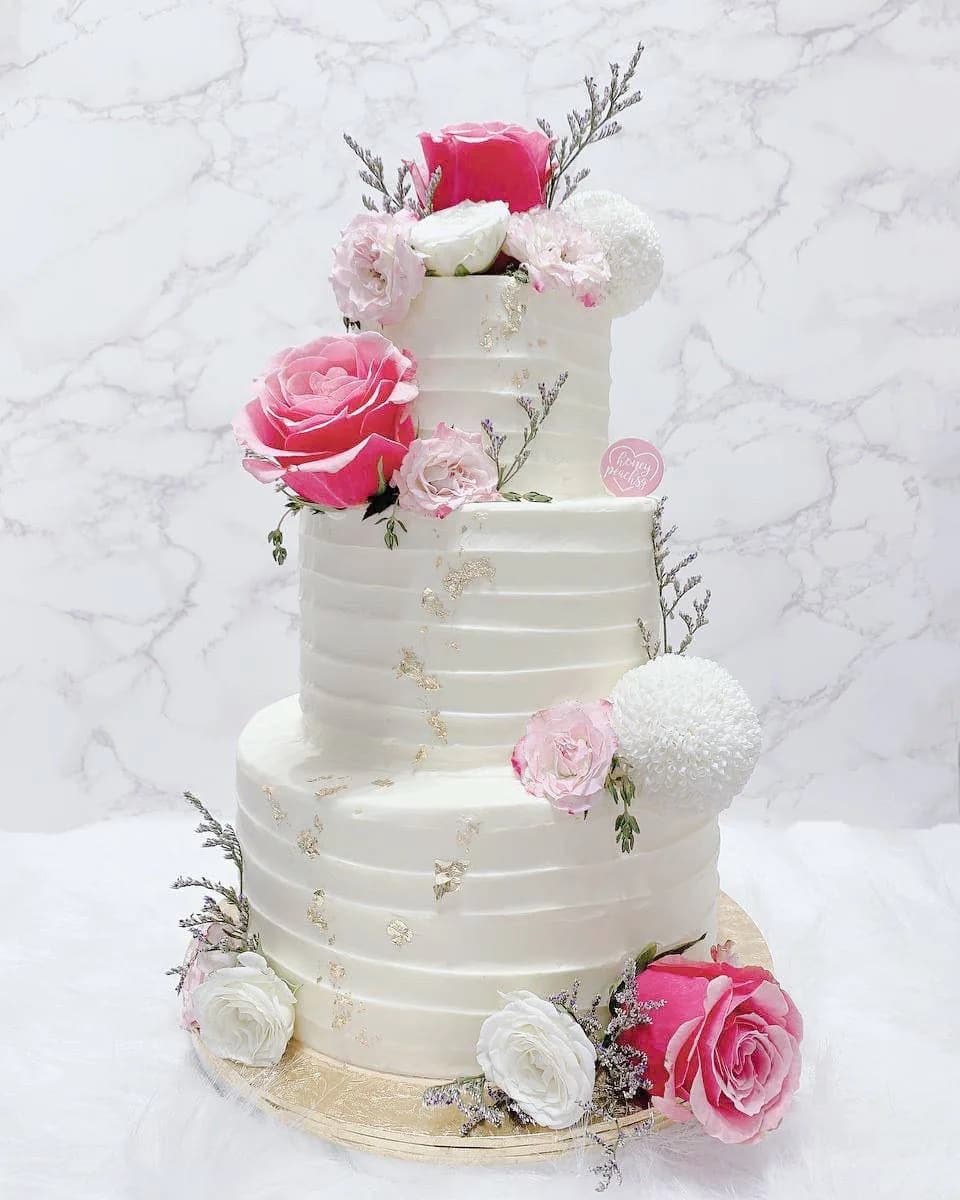 Four-tier wedding cake with white buttercream, fresh pink and red roses, white flowers, and gold details