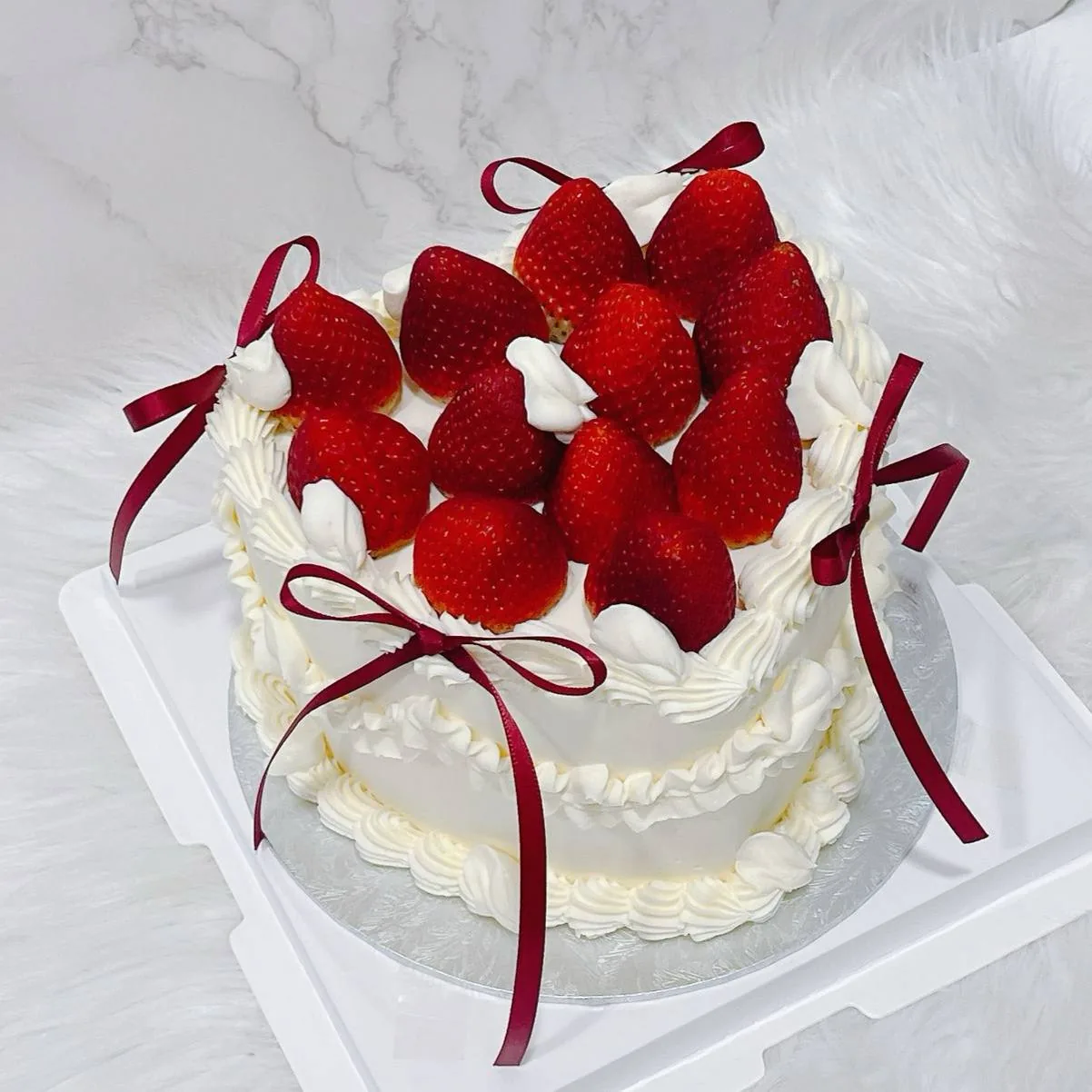 Round cake with white frosting, fresh strawberries, and burgundy ribbon decoration on white surface.
