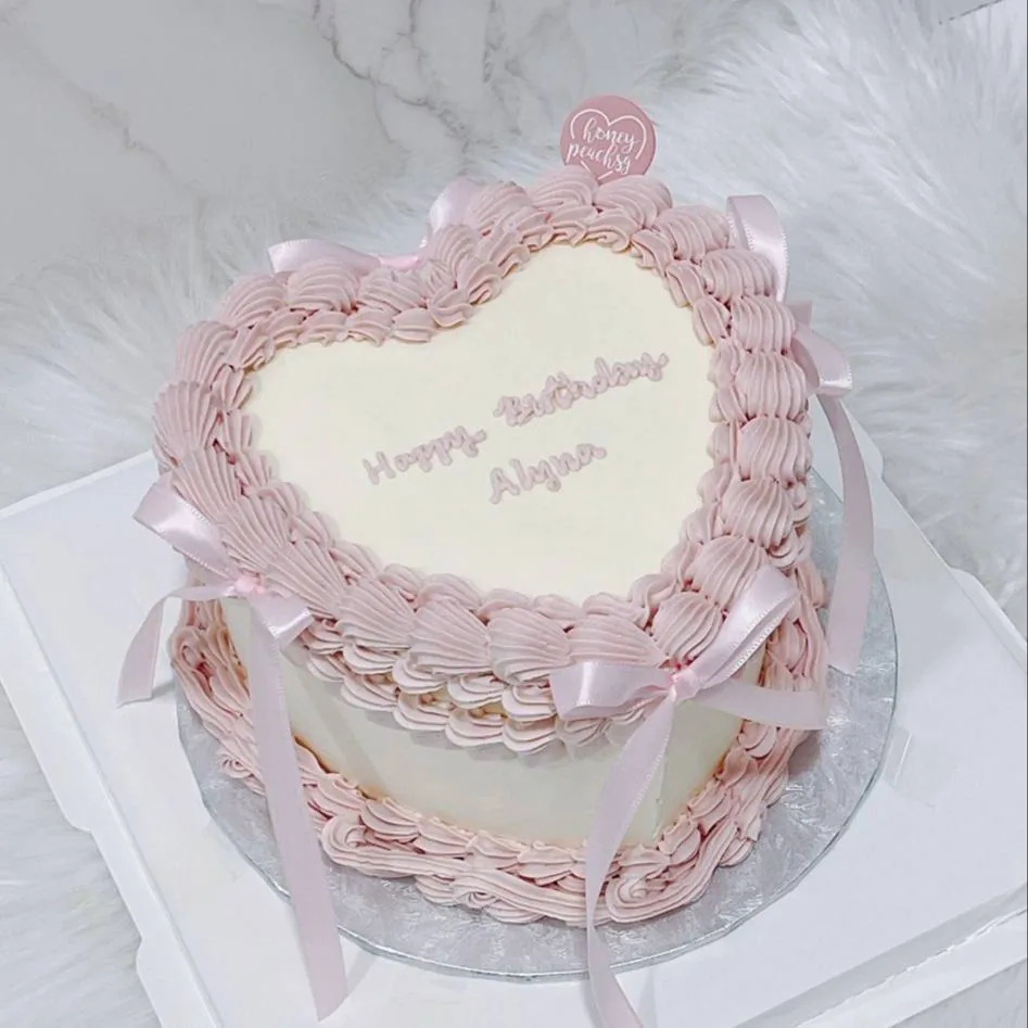 Two-tier heart-shaped cake with pink frosting piping, white fondant top, and pink ribbon bows