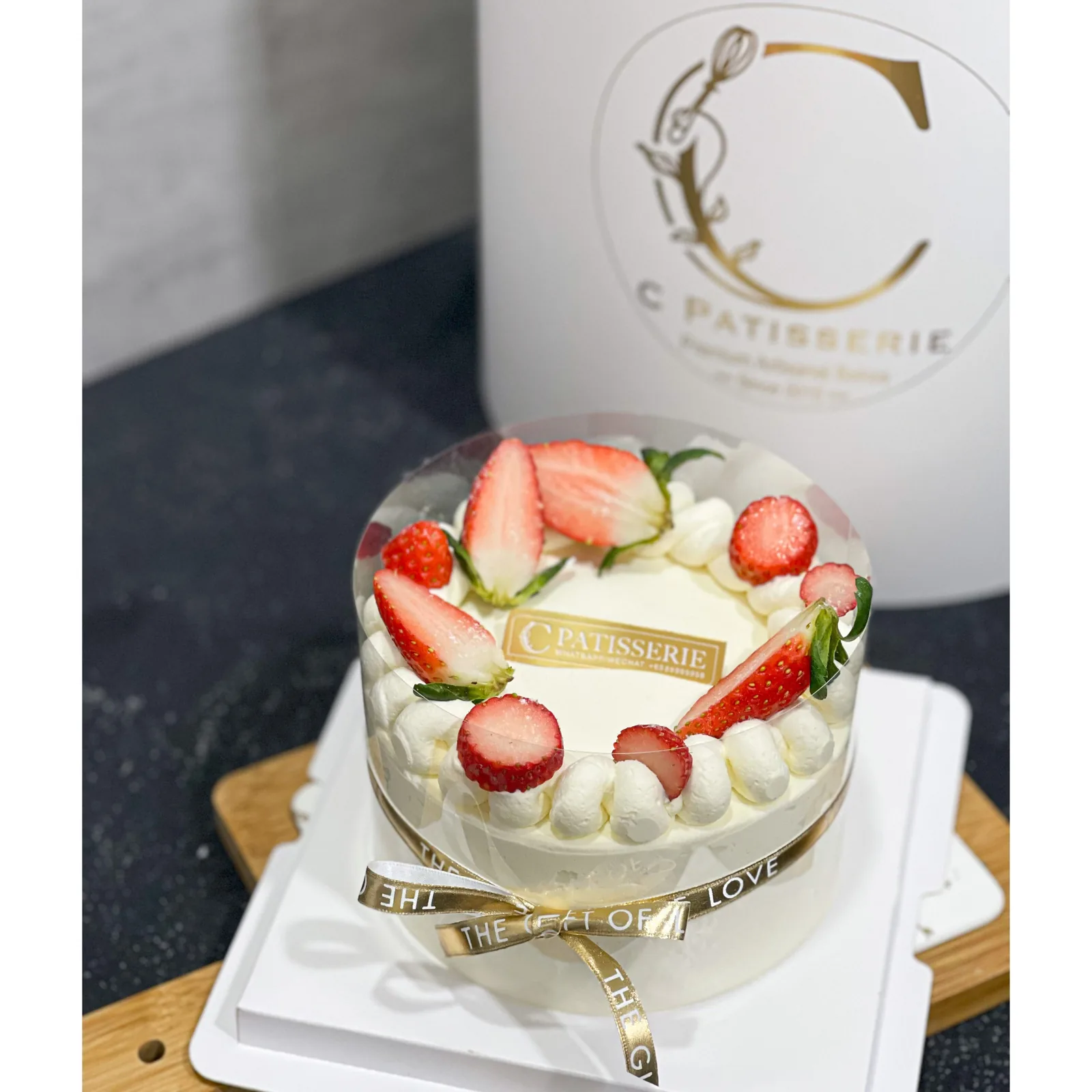 Strawberry cream cake with fresh berries, piped cream border, and gold ribbon, C Patisserie branding behind