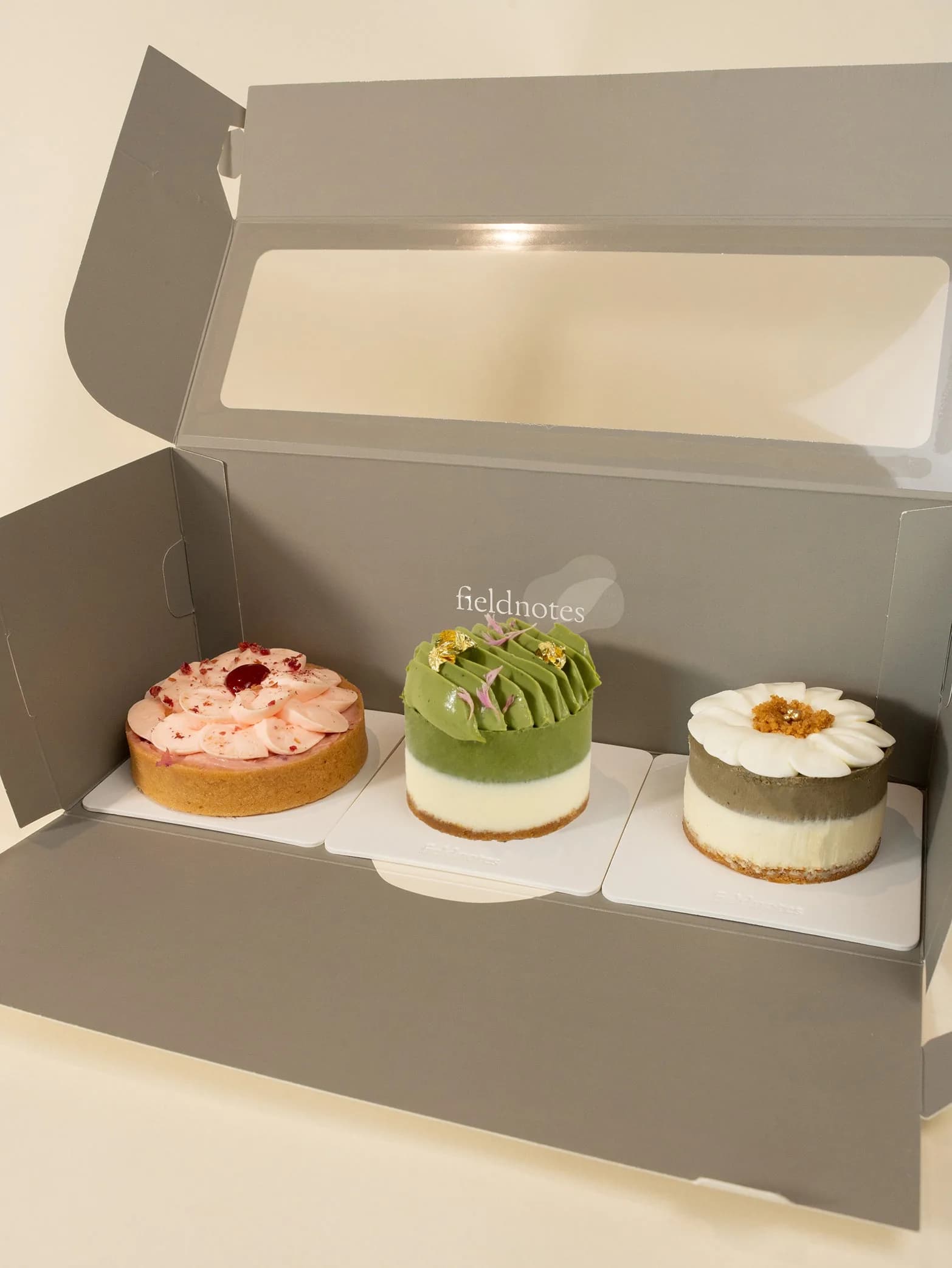 Three elegant layered cakes in shades of pink, green, and white displayed in branded takeout box