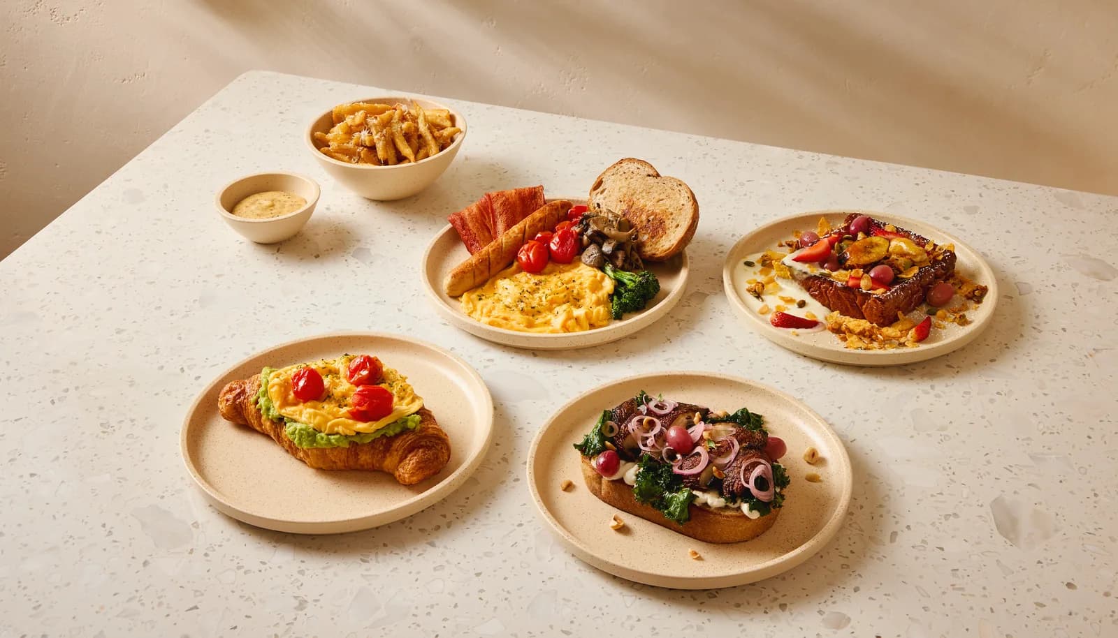 Breakfast and lunch dishes displayed on cream plates: egg croissants, loaded toasts, fries, and garnishes