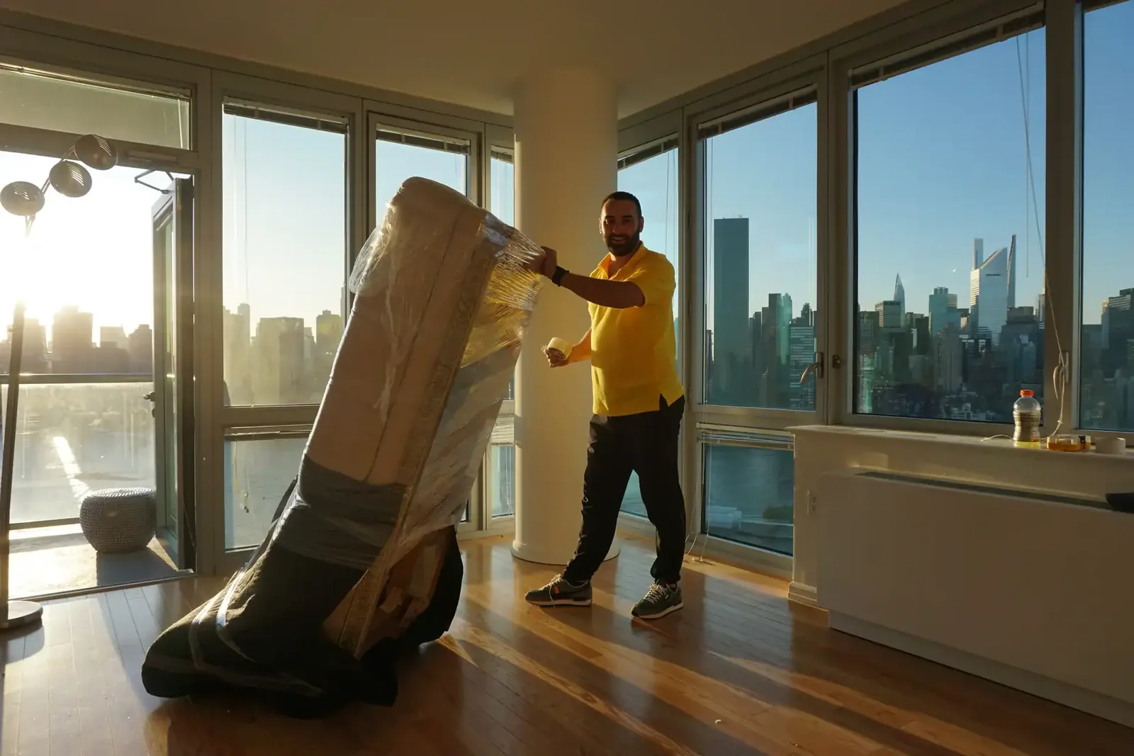 Mover wrapping furniture in a bright apartment with NYC skyline views through floor-to-ceiling windows