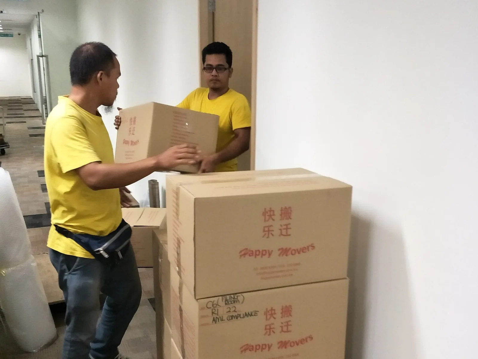 Two movers in yellow shirts carrying and stacking branded cardboard boxes indoors