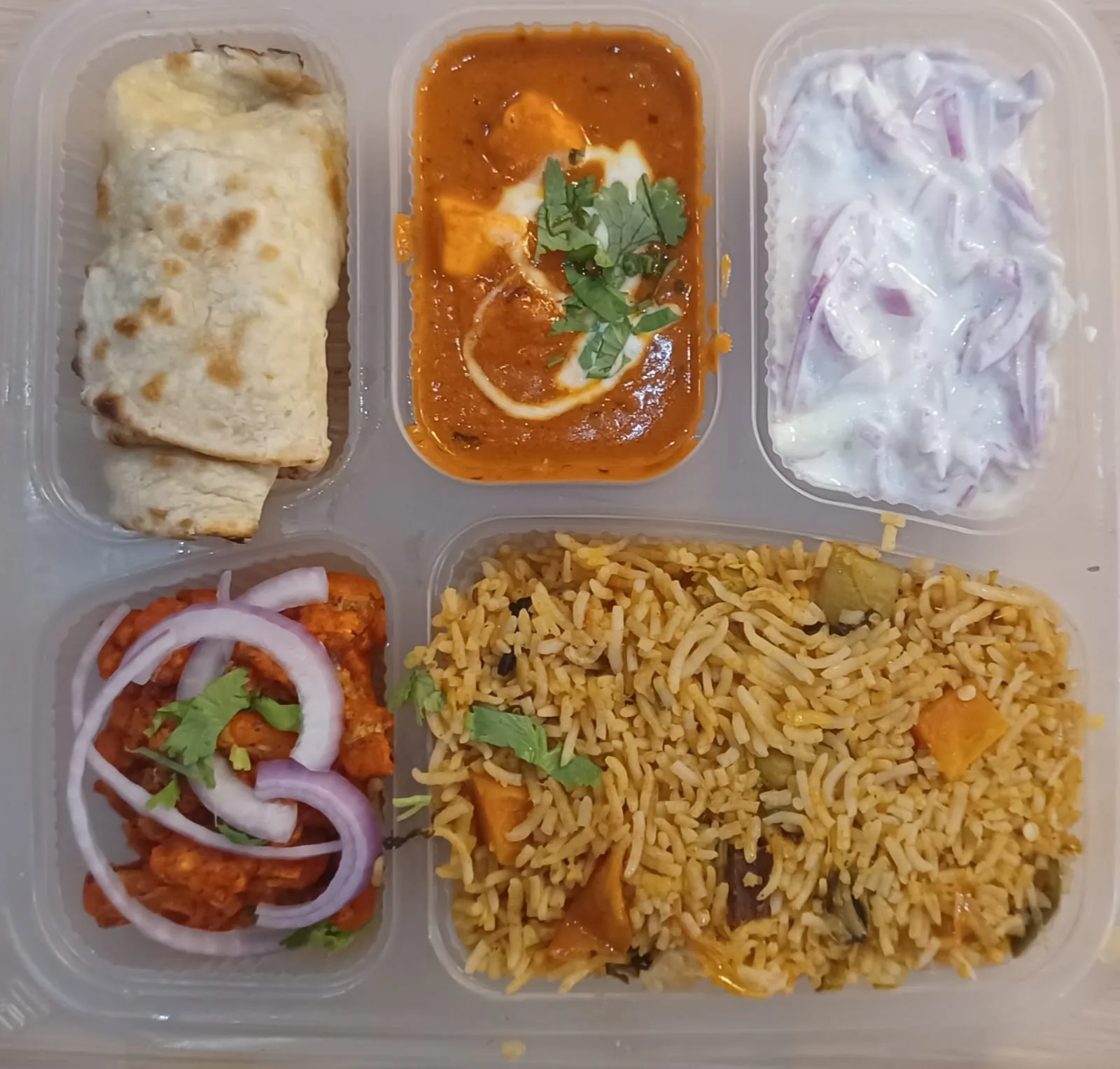 Compartmentalized meal tray with Indian dishes: wrapped item, curry, rice, raita, and paneer curry
