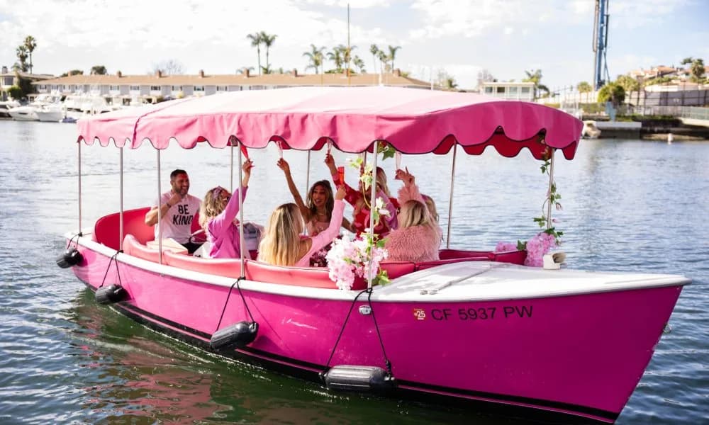 Pink party boat with passengers celebrating under a canopy on calm waterfront
