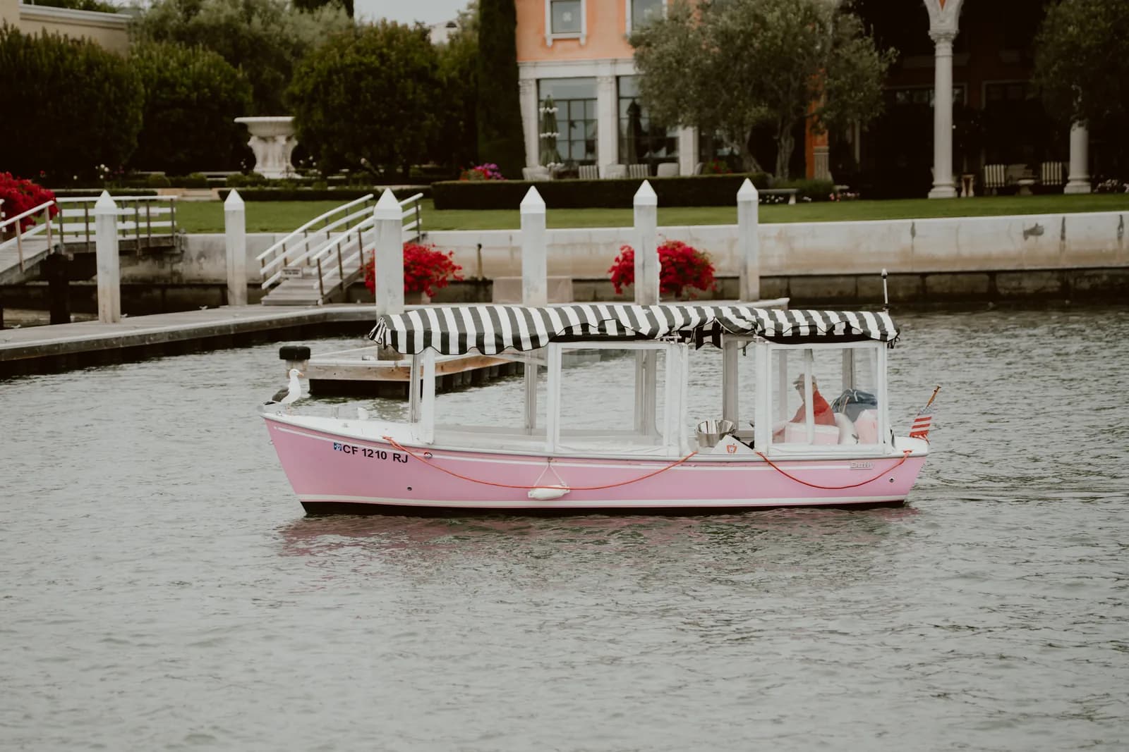 Pink party boat with striped canopy docked at waterfront estate with white railings and red flowers