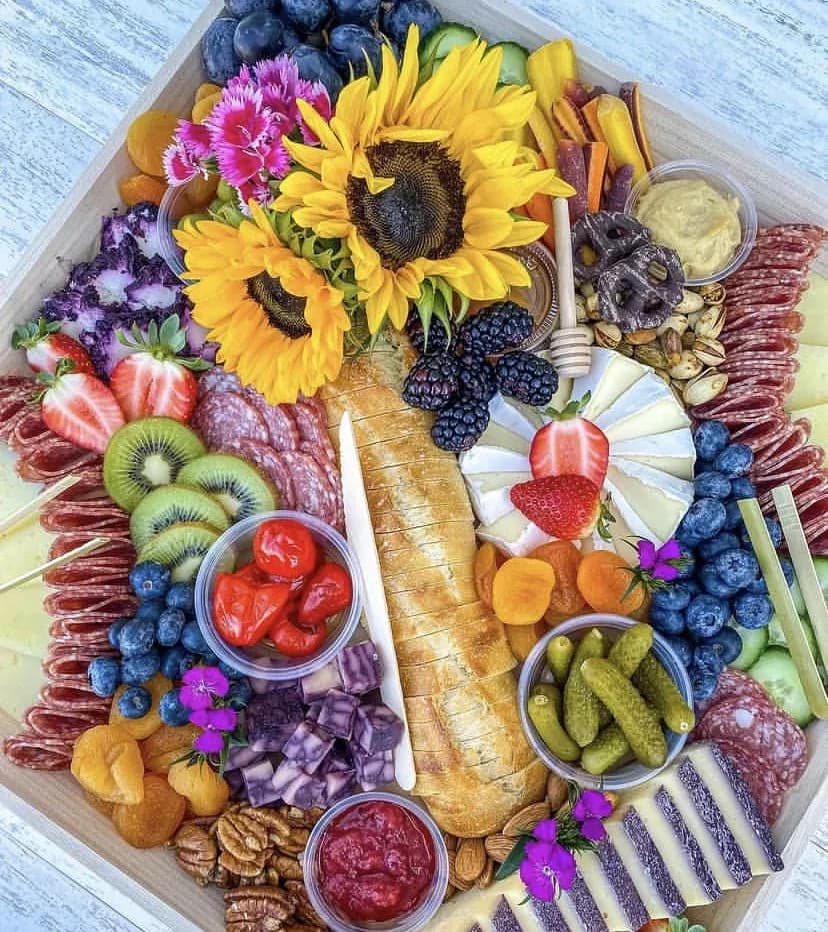 Elaborate charcuterie board with fresh produce, cured meats, cheeses, and sunflowers on white wood surface.